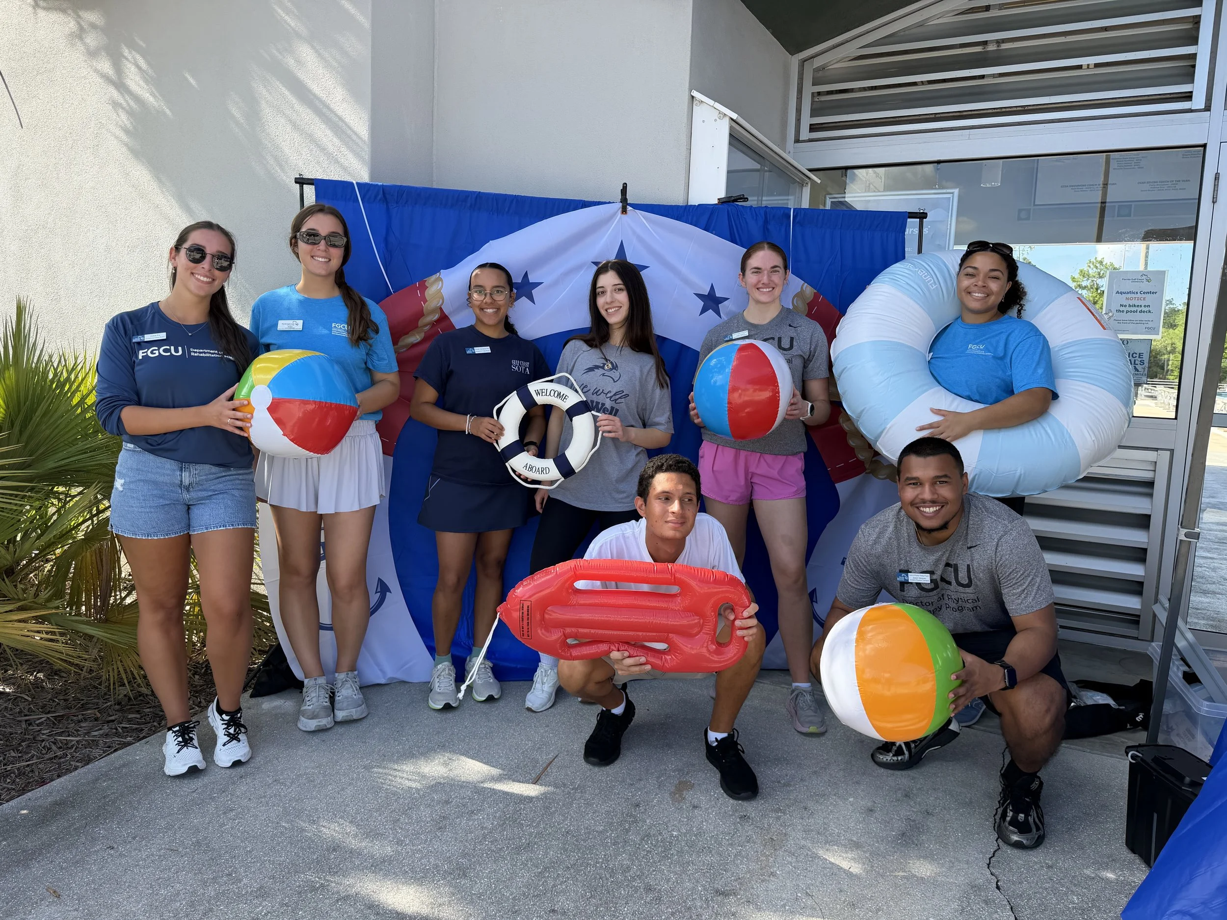 A group of six young adults, wearing casual and summer clothing, standing outside a building near a pool area, with some holding beach balls and a life preserver. They are smiling and appear to be enjoying a sunny day at a recreational or aquatic center.