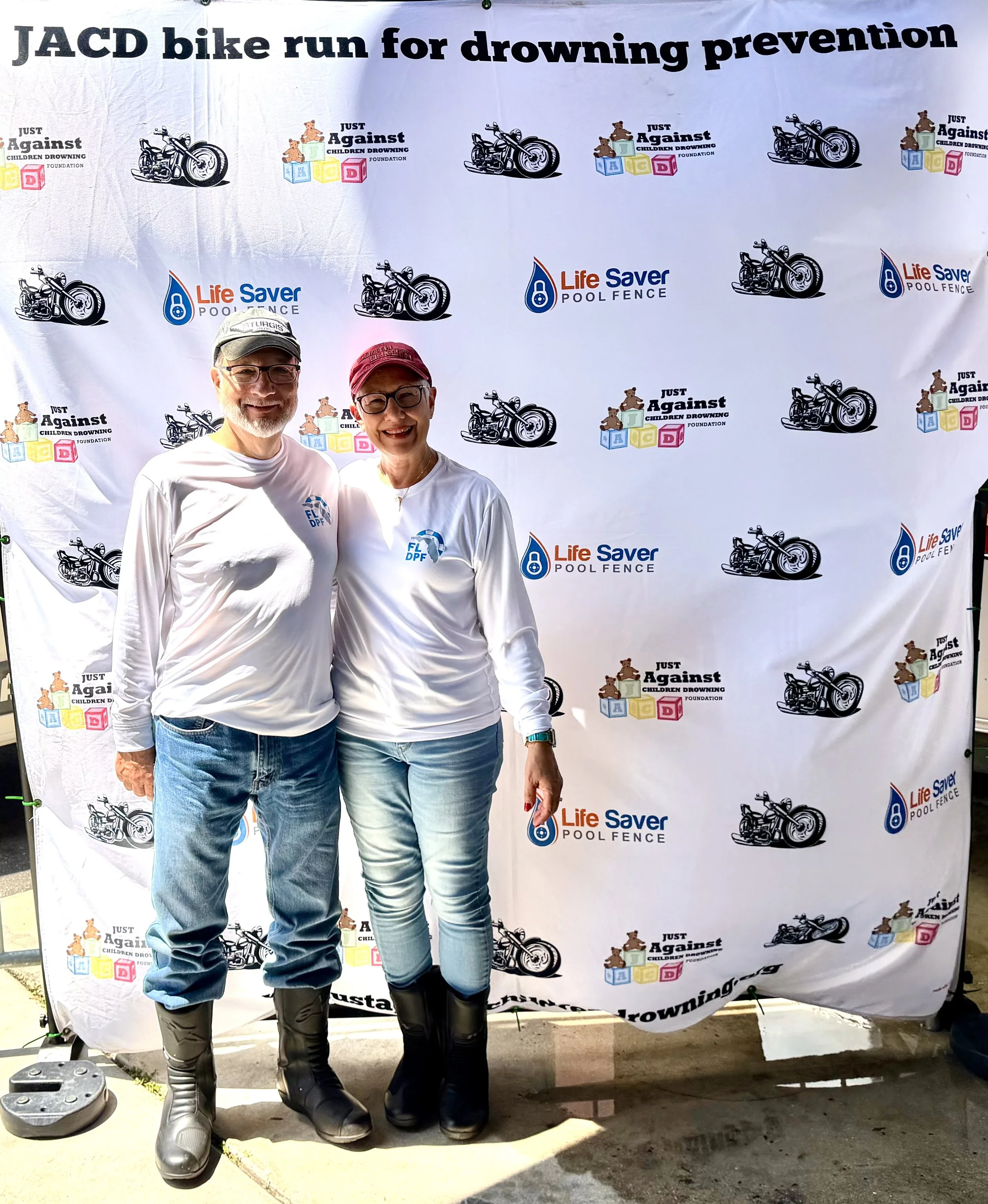 Two people standing in front of a backdrop with logos for a charity event. The man is wearing glasses, a baseball cap, a white long-sleeve shirt, and riding boots, smiling at the camera. The woman is wearing glasses, a red cap, a white shirt, and riding boots, also smiling at the camera.