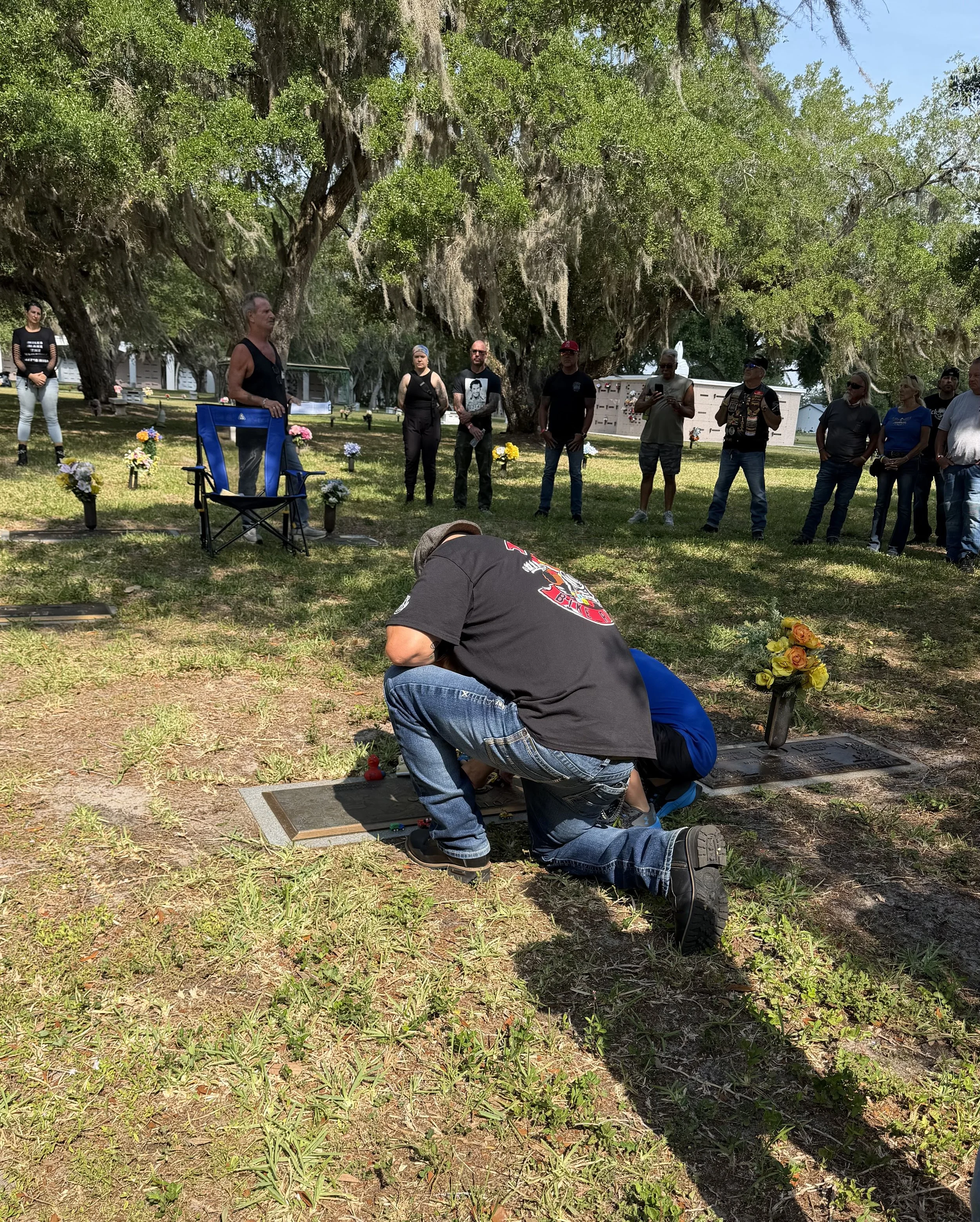 A group of people gathered outdoors on a sunny day near graves, with large trees in the background. Some individuals are standing, one is kneeling and tending to a grave, and others are observing. Flowers are placed at some graves.