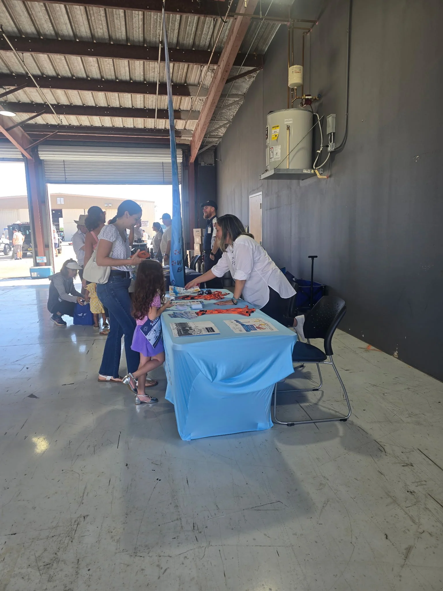 People gathered around a table with a blue tablecloth in a warehouse-like space, engaging in an event or activity. There are chairs and various informational materials on the table.