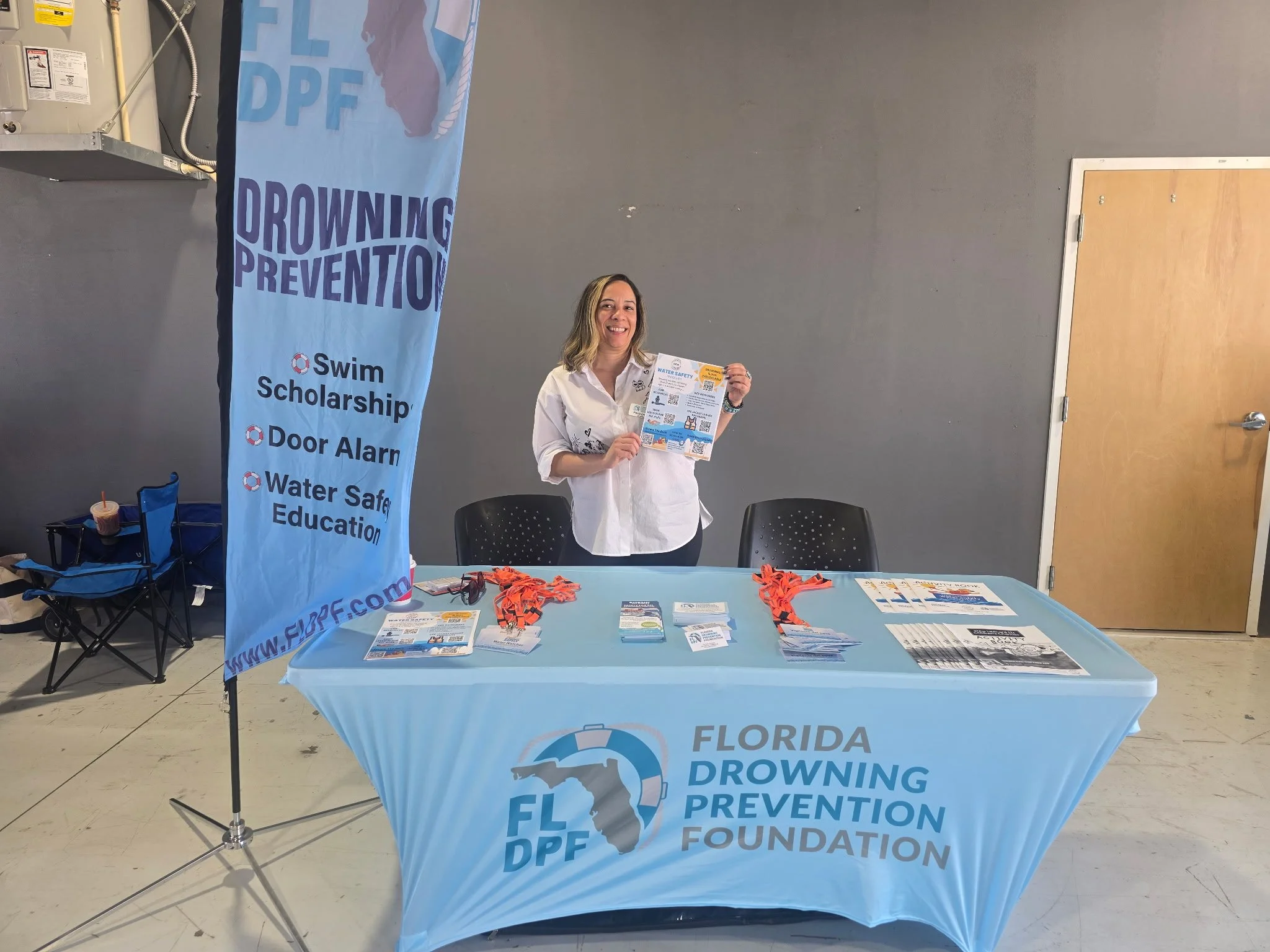 A woman standing behind a table at a Florida drowning prevention event holding a flyer. The table has bookmarks and informational materials. A blue banner with drowning prevention messages and a Florida map graphic is beside her.