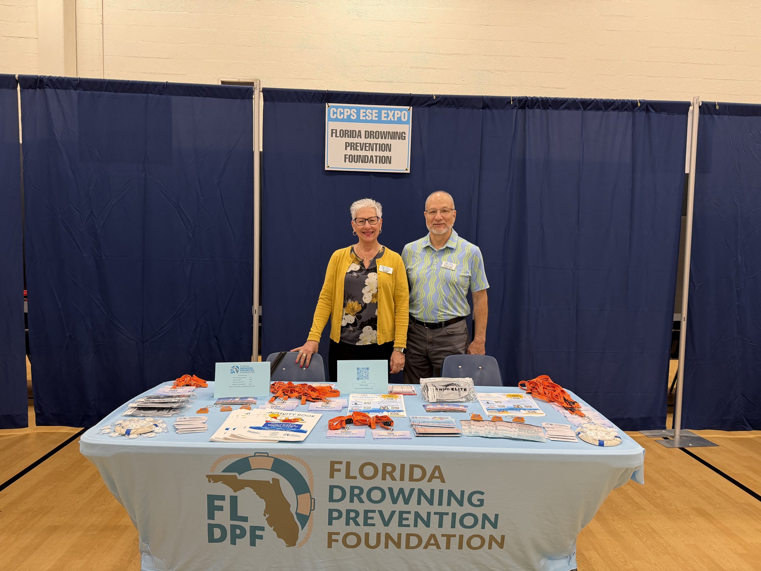 Two people standing behind a table with Florida Drowning Prevention Foundation promotional materials at an expo. The table has a Florida-shaped logo and the people are smiling. The background shows a navy blue privacy screen with a sign reading 'CCPS ESE Expo Florida Drowning Prevention Foundation.'