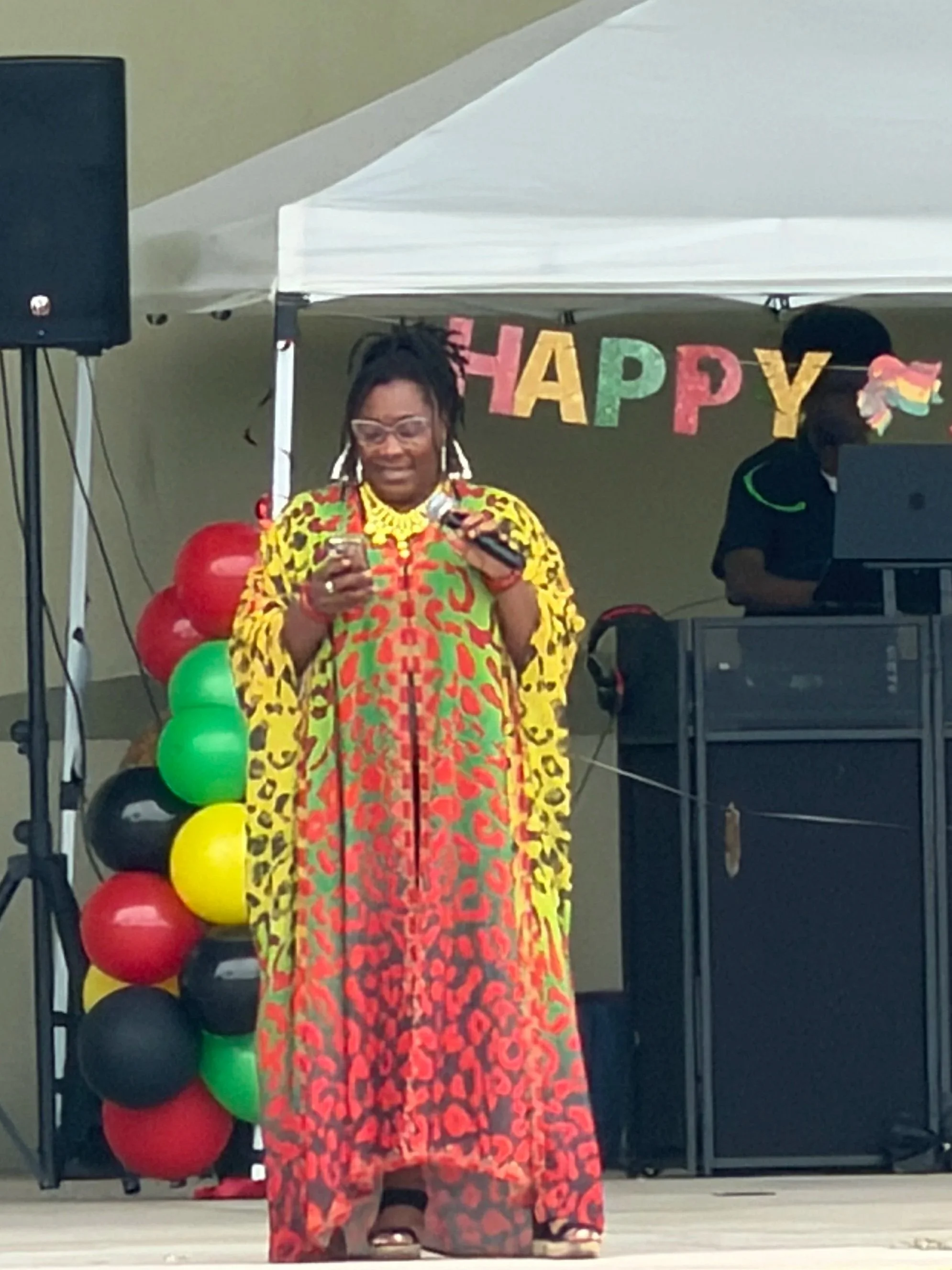Woman in colorful dress speaking into a microphone at a celebration with balloons and a 'Happy' banner in background.