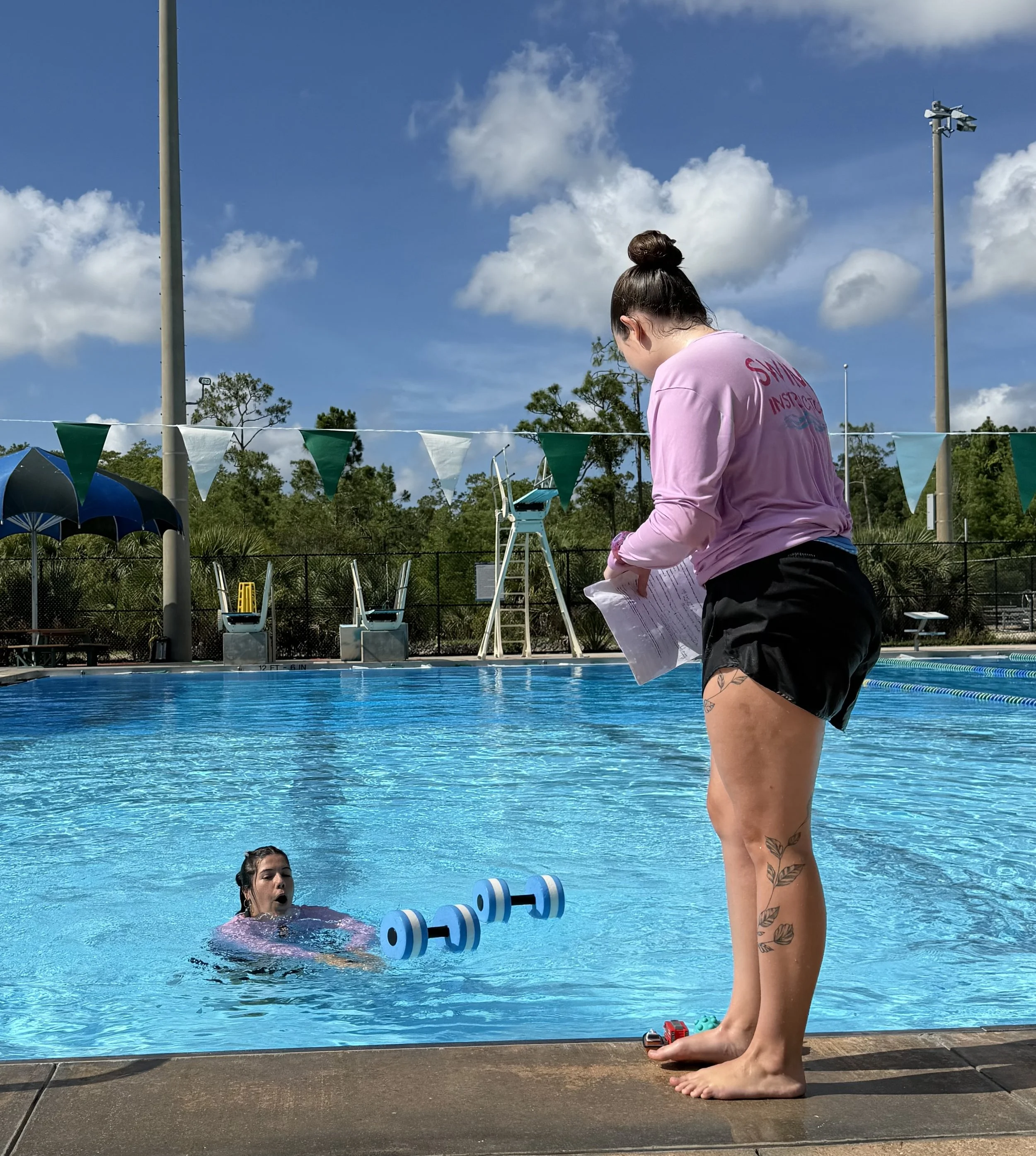 A swimming pool scene with two people. One person is swimming in the pool, and the other person, standing barefoot on the pool's edge, is holding papers and looking down. There are swimming lane dividers in the pool, umbrellas and a scoreboard in the background, along with trees and blue sky with clouds.
