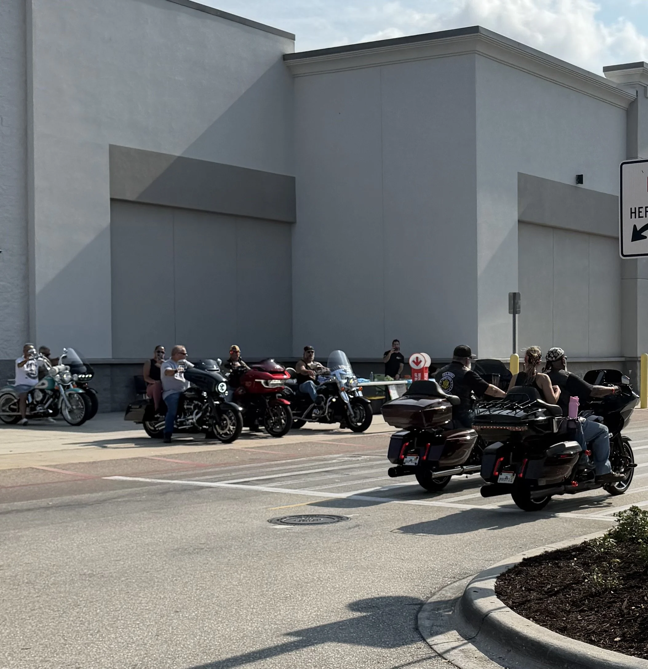 A group of motorcyclists and people standing outside a building with a white exterior wall, partly visible sign on the right, a sidewalk, and a parking lot with multiple motorcycles parked.