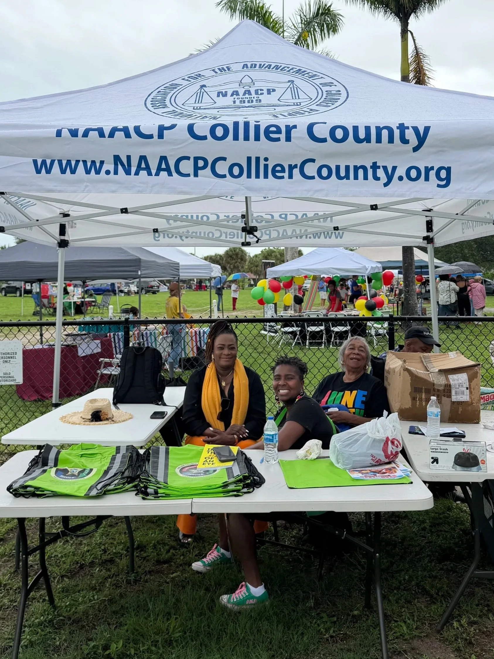 People sitting at a table under a NAACP tent at an outdoor event, with various items like shirts, flyers, and water bottles on the table, and a grassy field with additional tents and balloons in the background.