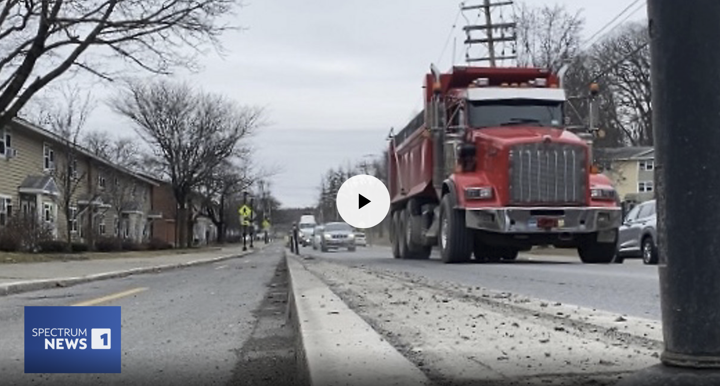 A red truck driving on a residential street with trees, houses, and parked cars, captured from a low angle near the curb, with a Spectrum News 1 logo in the bottom left corner.