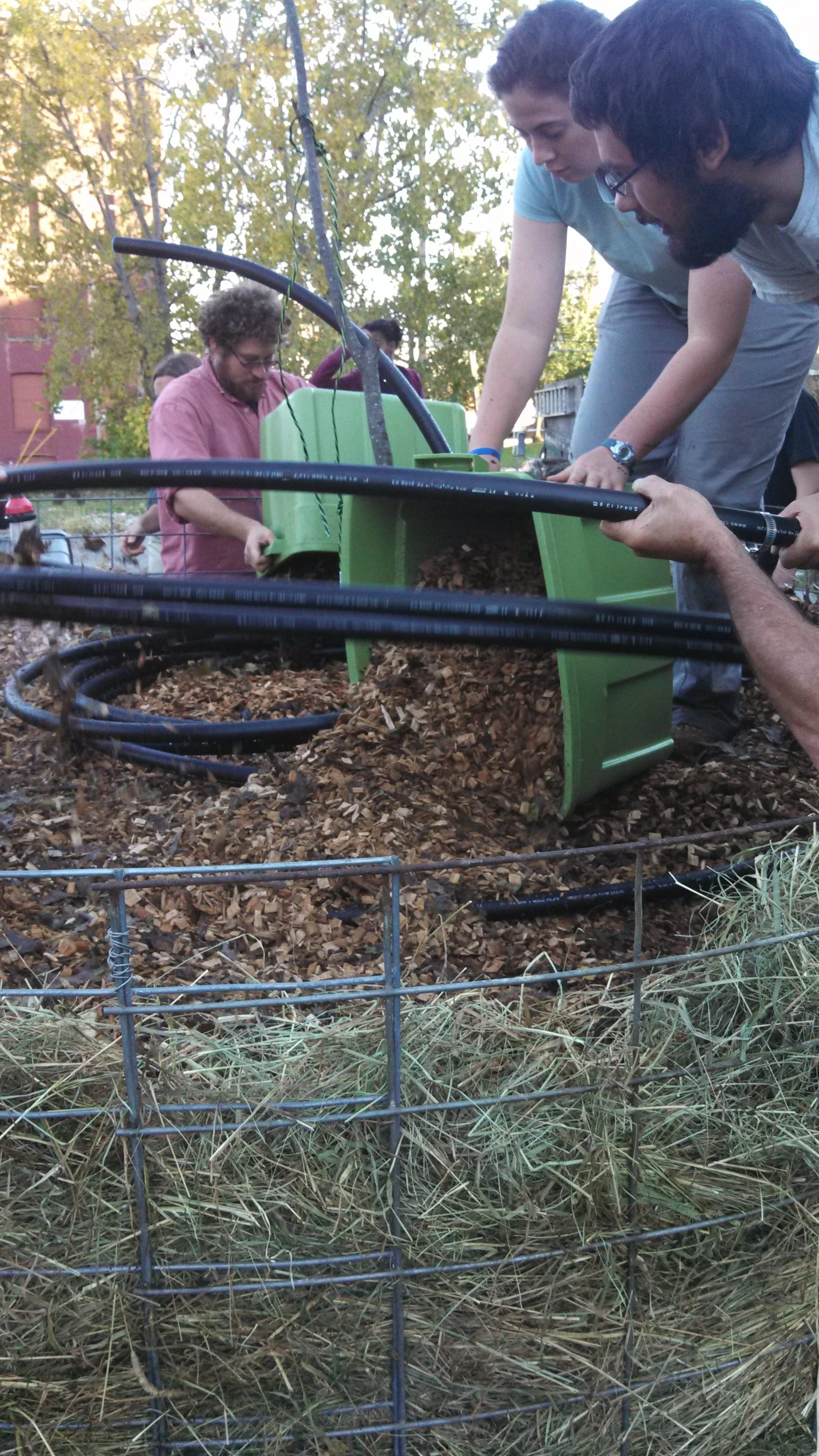People using a green compost bin to turn wood chips, outdoors with trees.