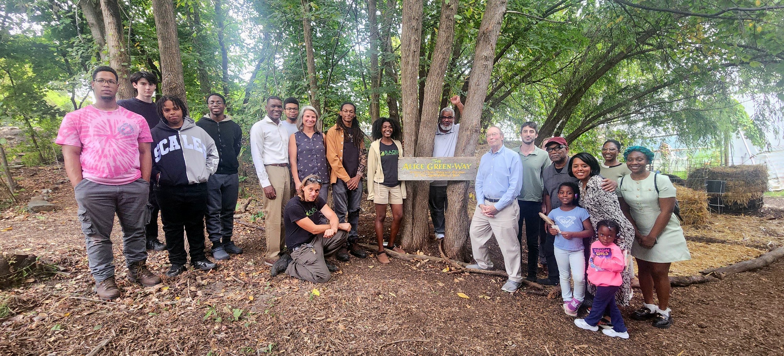 Group of people standing outdoors in front of large trees, some smiling, with a wooden sign that reads 'The ALICE GREEN-WAY'.