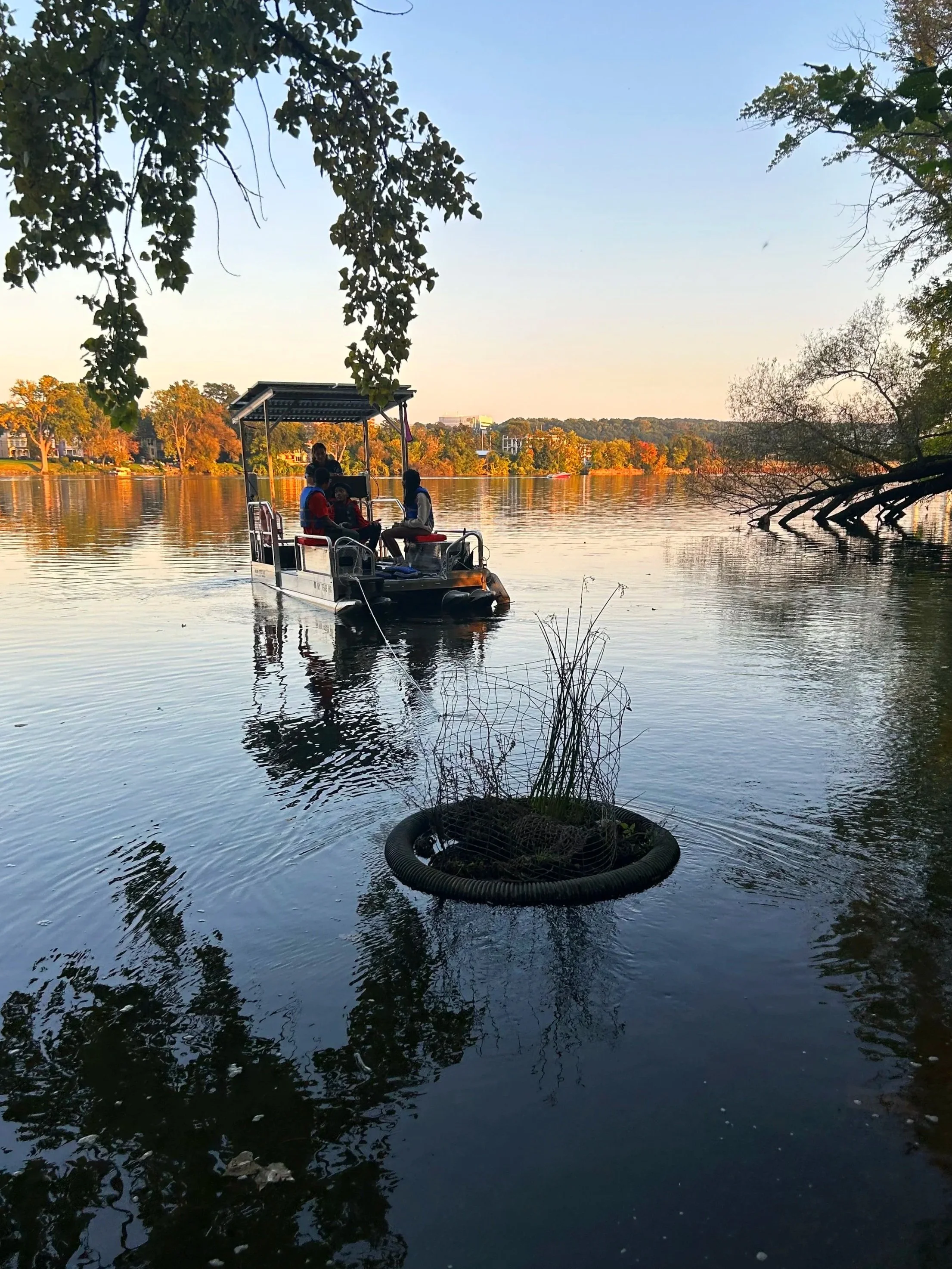 A boat with people on the Hudson River, surrounded by trees with fall foliage, during sunset.