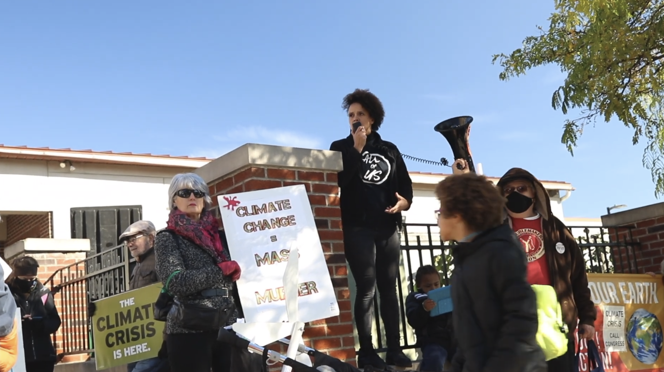 People gathered at a climate change protest outdoors; a woman with curly hair stands on a brick platform speaking into a megaphone, surrounded by signs about the climate crisis.