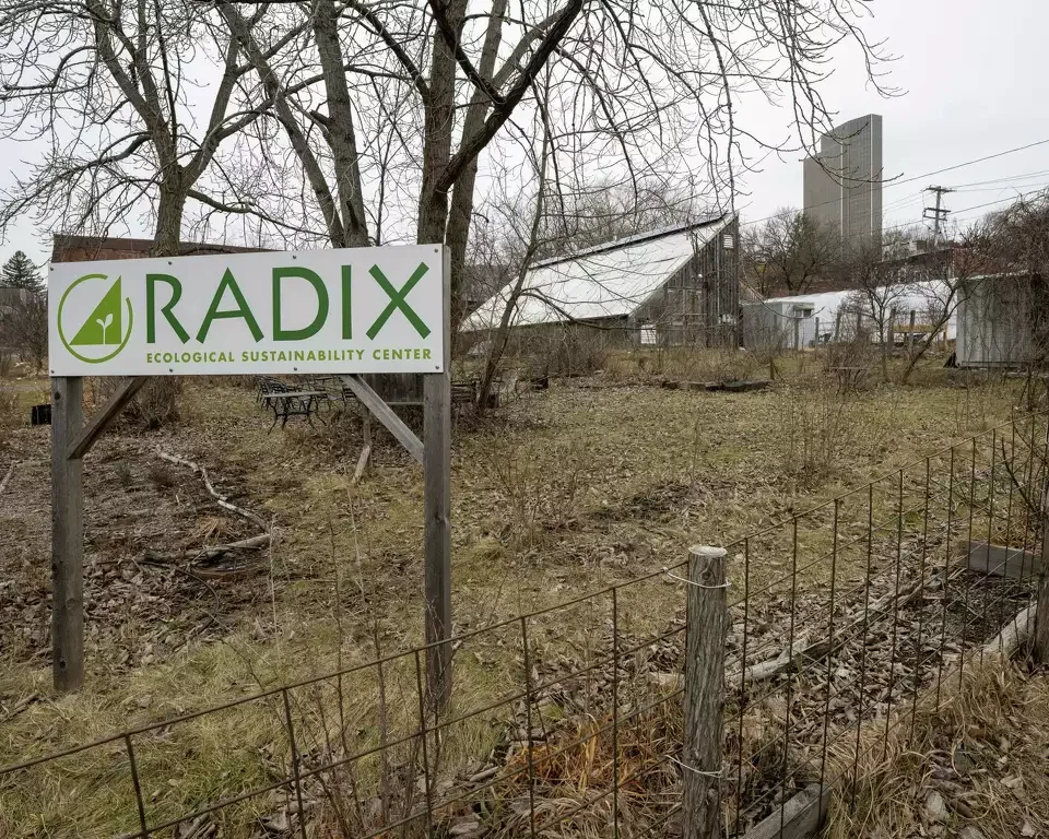 Sign reading 'RADIX Ecological Sustainability Center' in front of an outdoor area with sparse grass, leafless trees, and a makeshift greenhouse structure, with a fence and some buildings in the background.