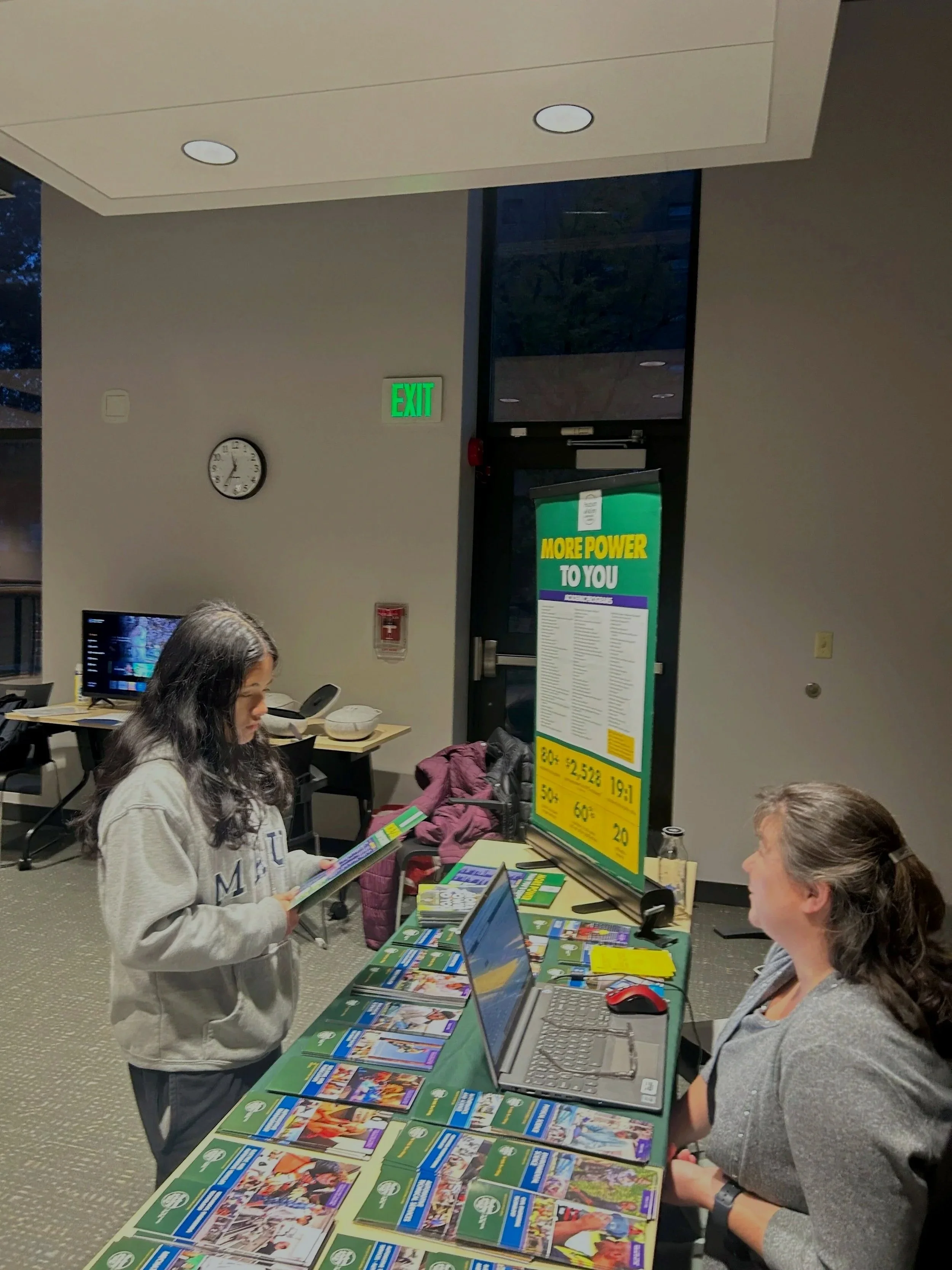 A woman sitting at a table with a laptop, talking to a girl standing across from her, who is holding a book. The table has many brochures or flyers, and there is a large informational poster on the wall behind them.