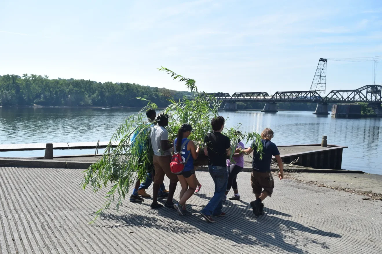 Group of people walking along river bank with a large plant, with an industrial bridge and water in the background on a sunny day.