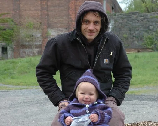 A man with dark hair and a black jacket smiling behind a toddler in a purple hoodie, sitting outdoors on a gravel path with greenery and a brick building in the background.