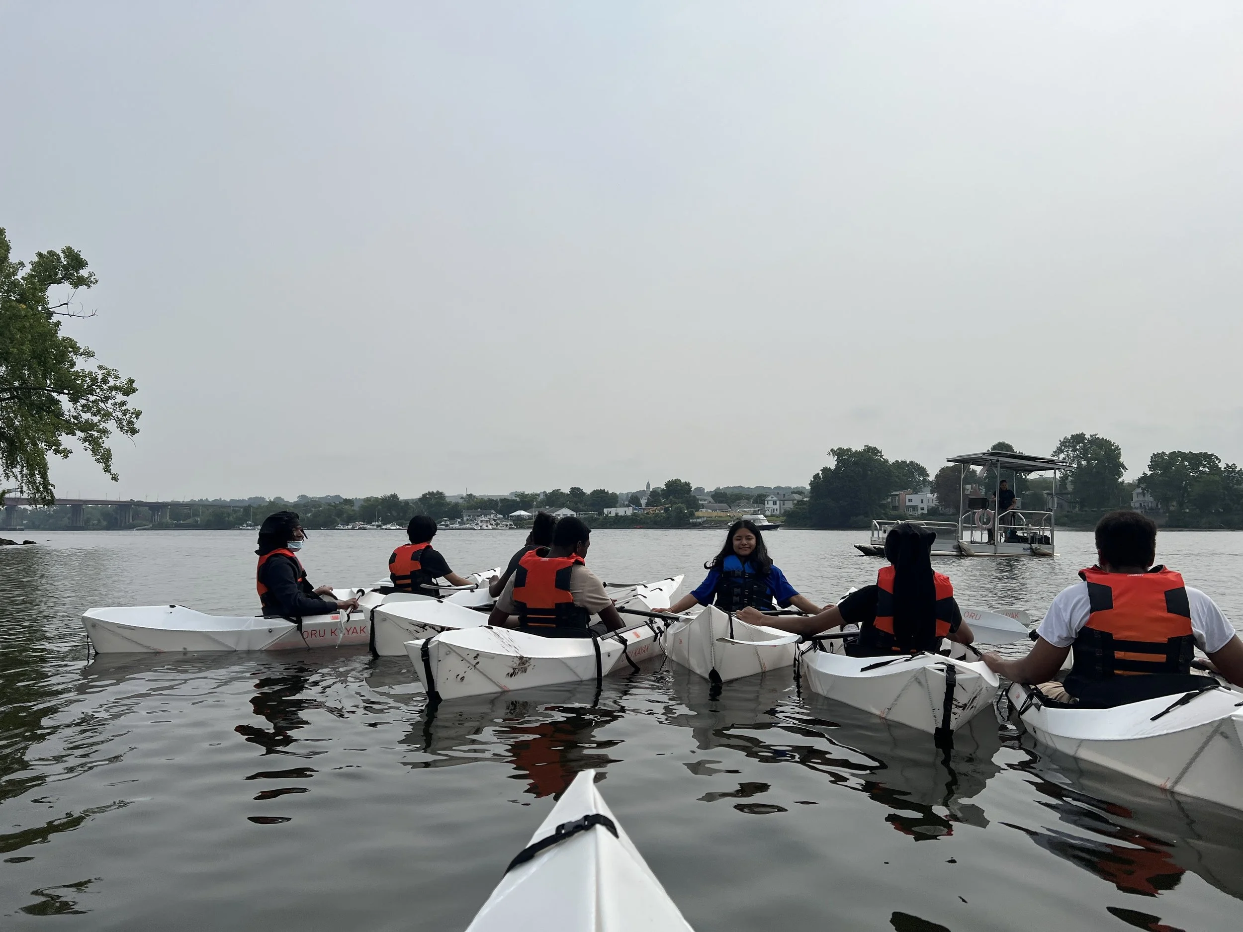 Group of people kayaking on a calm river, with some wearing life jackets and a small boat in the background.