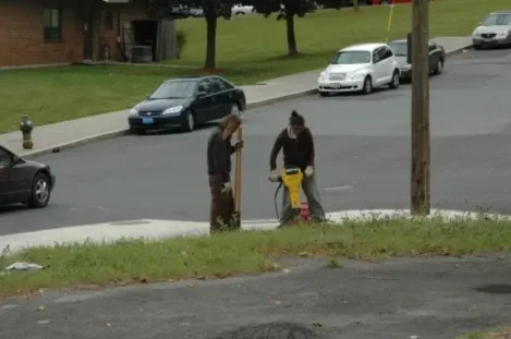Two people jackhammering on a grassy area near the street.