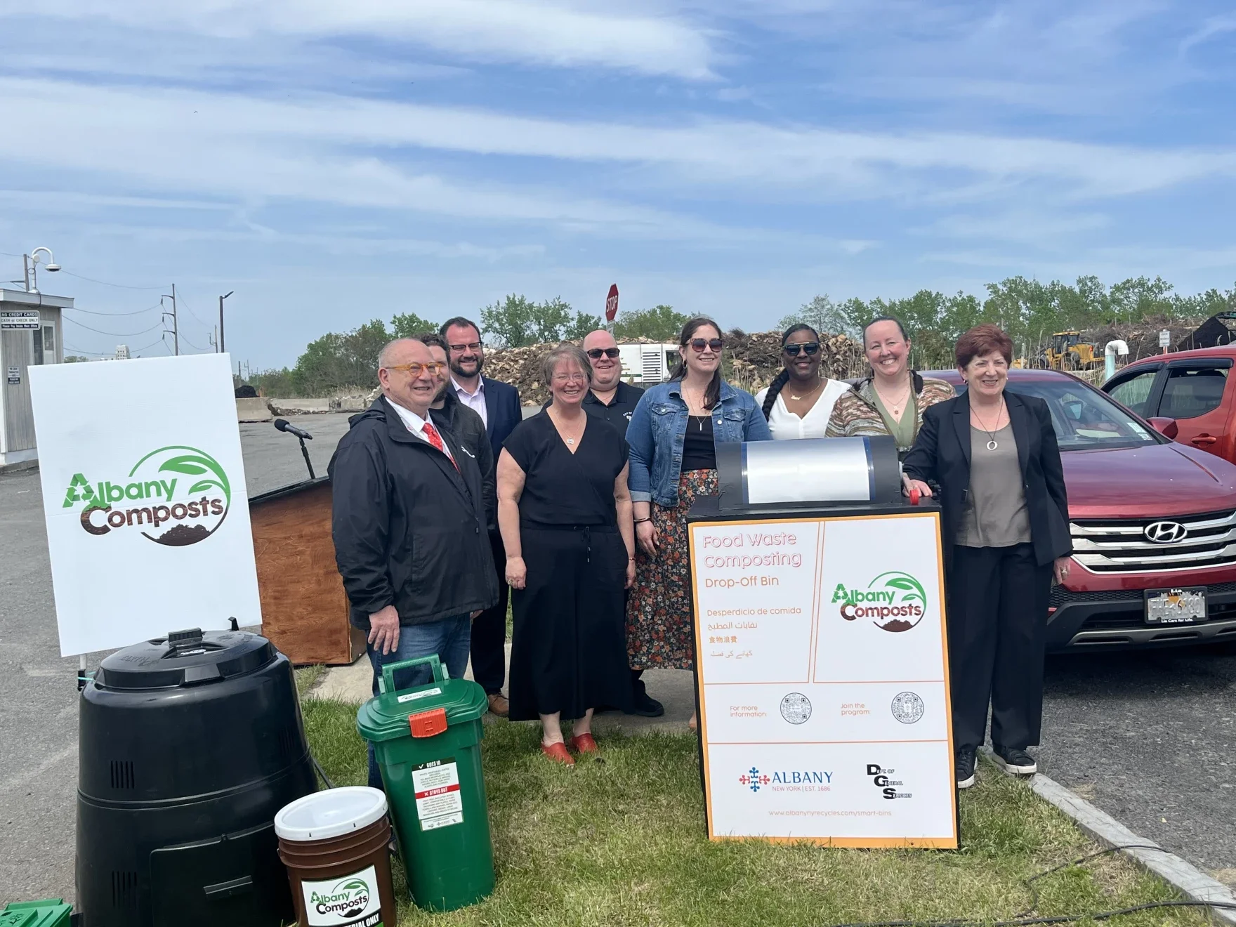 Group of people standing outdoors near a food waste composting drop-off bin with signs for Albany Composts, cars in the background, and a cloudy sky.