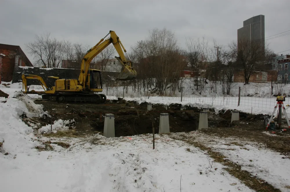 A construction site in winter with a yellow excavator digging a hole, concrete pillars in the ground, and a tripod with surveying equipment, with snow-covered ground and bare trees in the background.