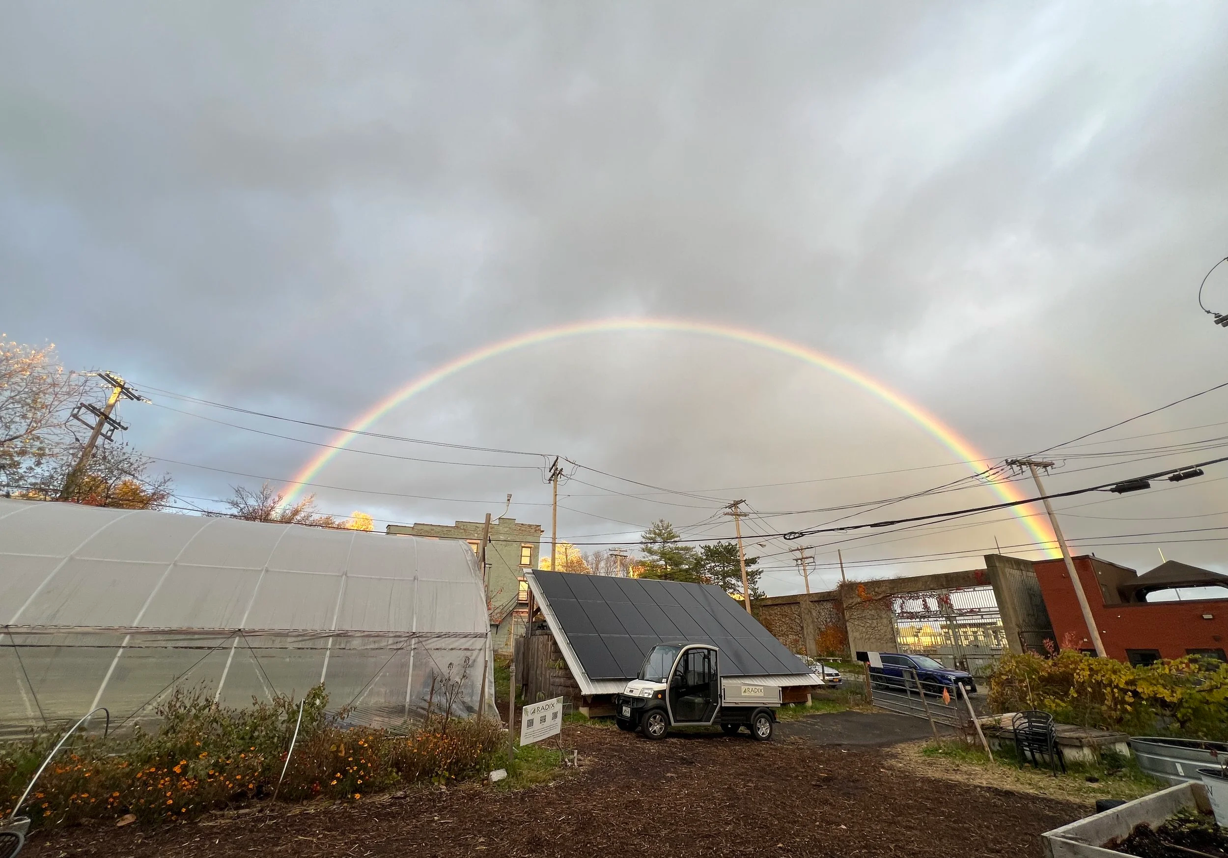 A rainbow arching over the garden with a hoop house, solar panels, and houses under a cloudy sky.