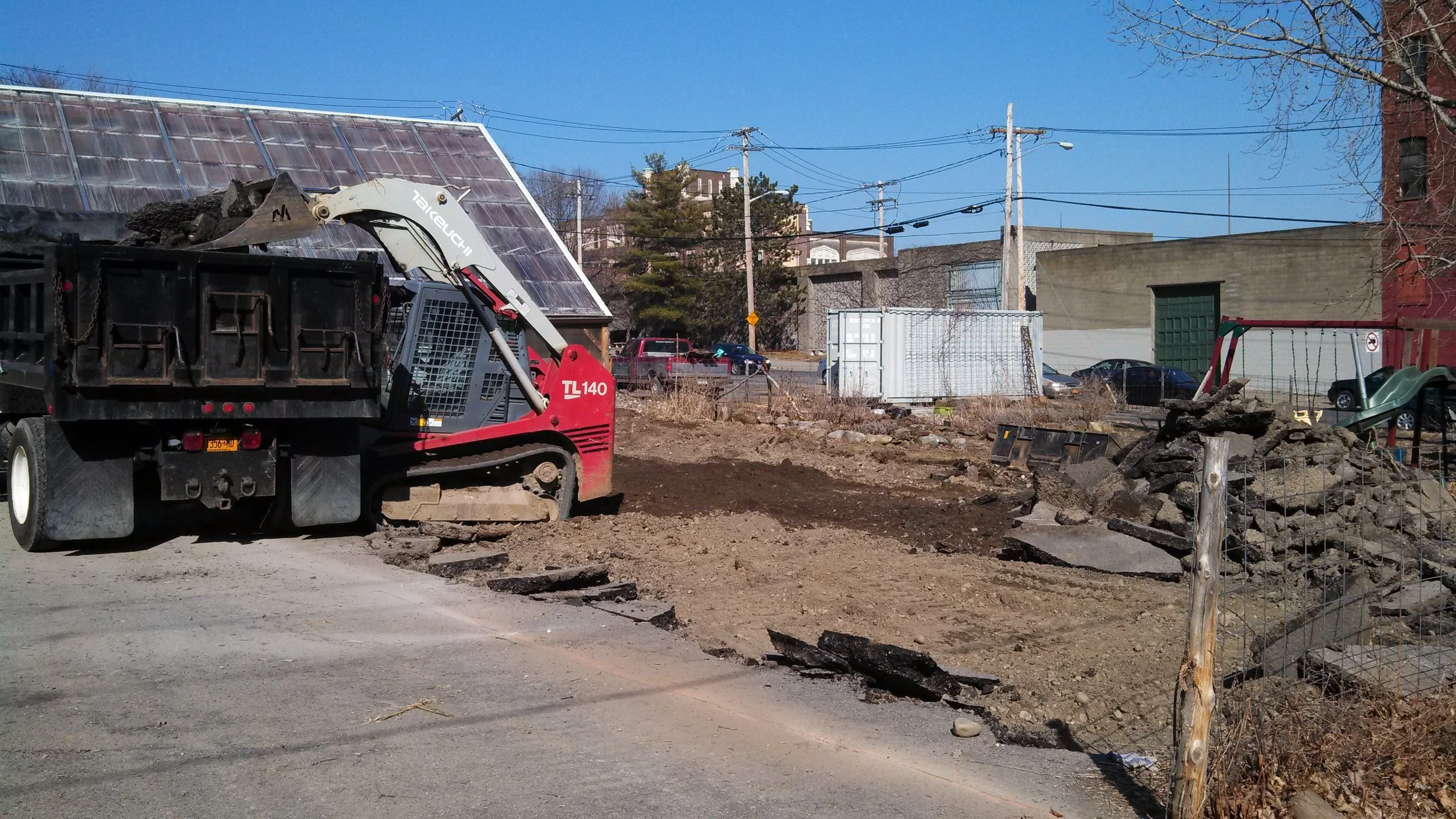Construction site with an excavator removing dirt and rocks, debris piled to the right, on a sunny day with a clear blue sky, residential buildings, parked cars, and utility poles in the background.