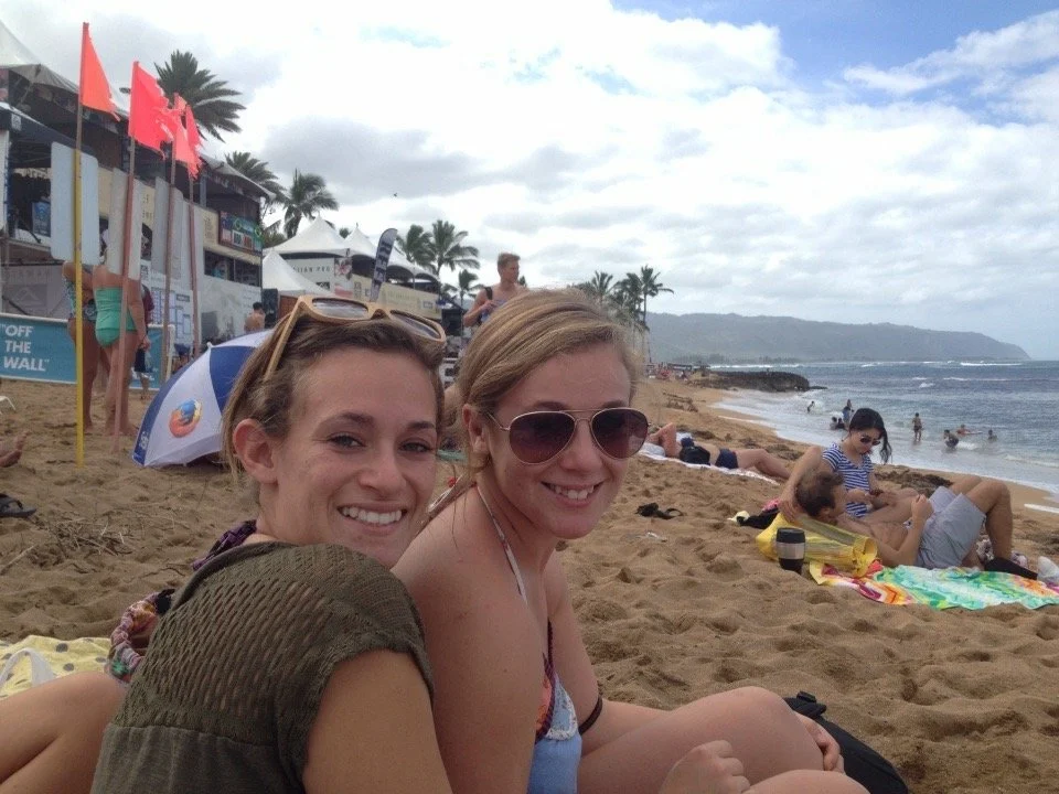 Two women sitting on the sand at a beach, smiling at the camera, with people, umbrellas, and palm trees in the background.
