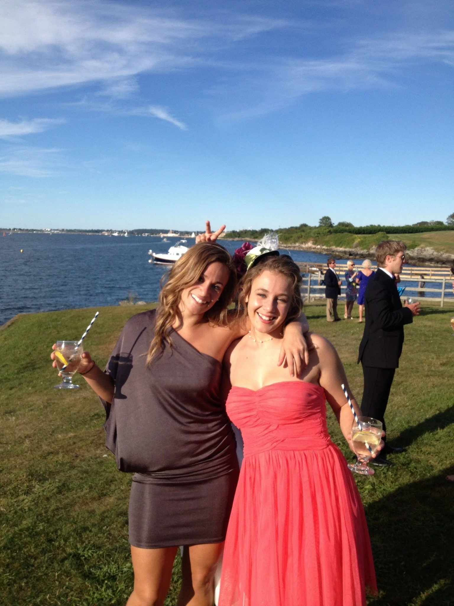 Two young women in dresses holding drinks and smiling at an outdoor event near water, with other people in the background.