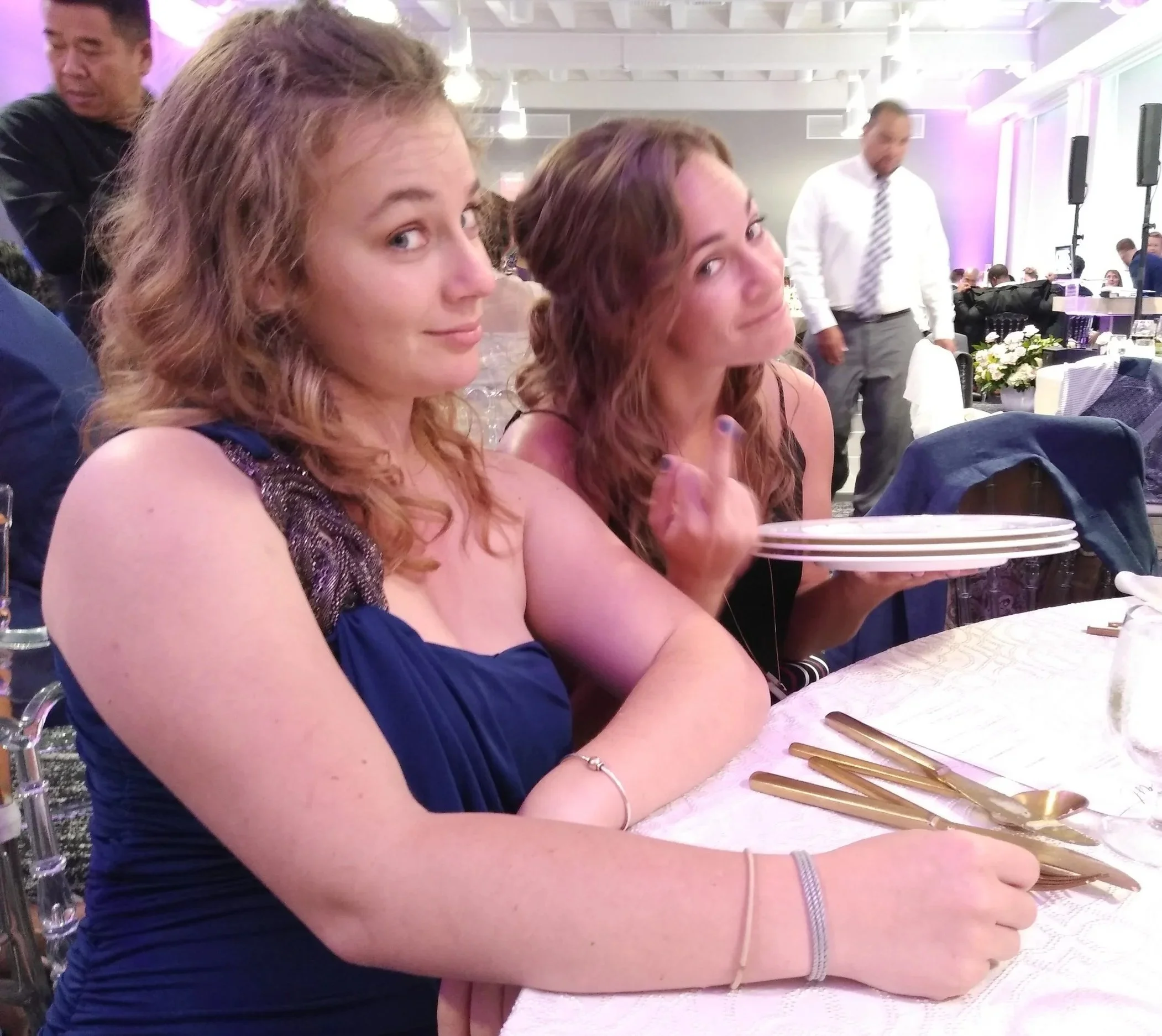 Two women with curly hair sitting at a formal event, one in a dark blue dress and the other in a black dress, with a tray of plates in her hand. The woman on the right is flipping the middle finger and smiling.