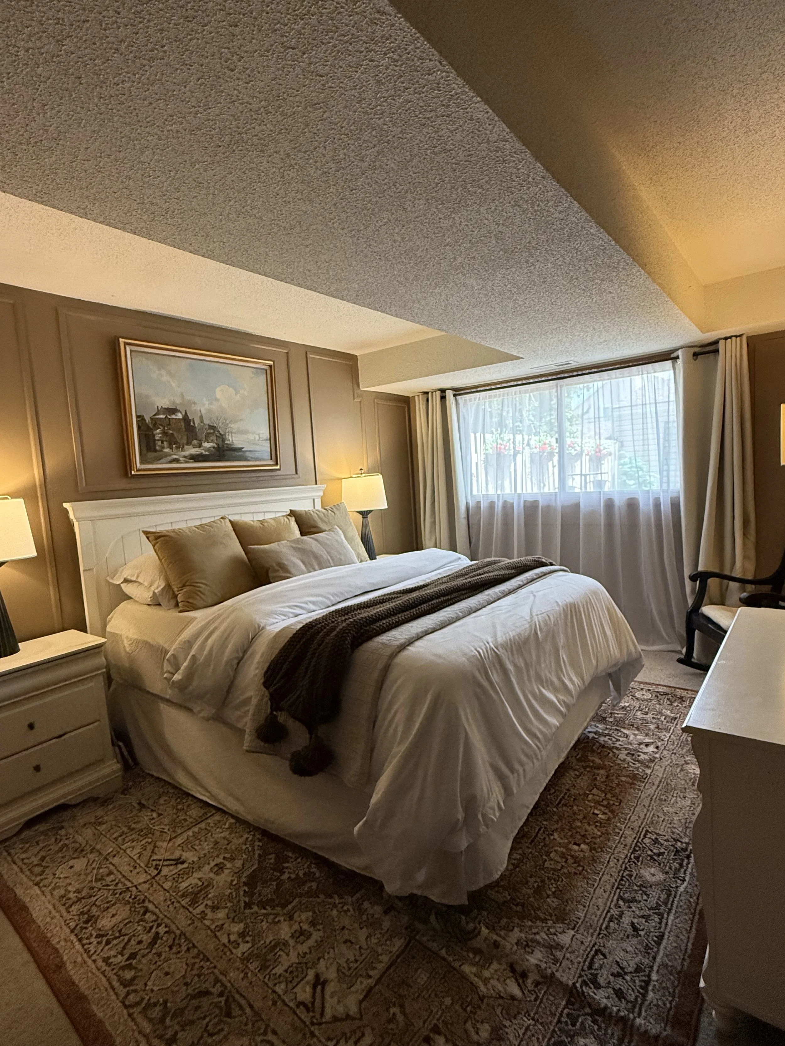 A cozy bedroom with a large bed that has multiple pillows and a white headboard. There is a nightstand with a lamp on each side of the bed. The room features a carpeted floor, a rug, a window with curtains, and a painting hanging above the headboard.