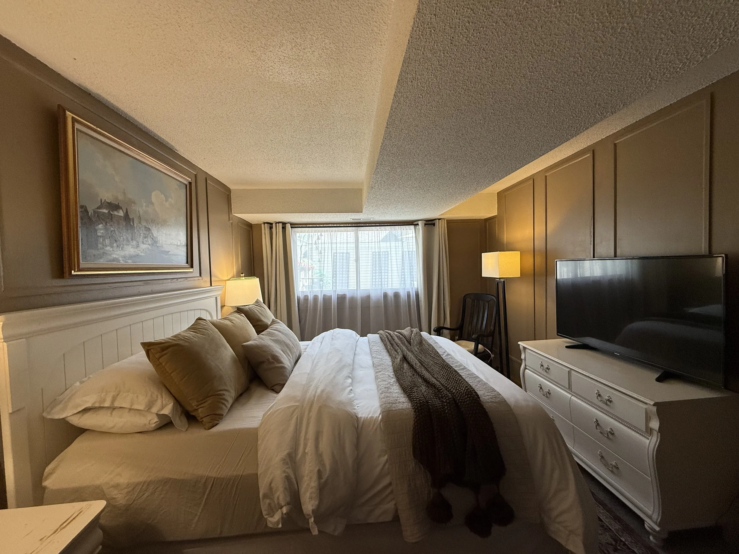 Bedroom with a white bed, beige pillows, a dresser with a TV, and a window with curtains.