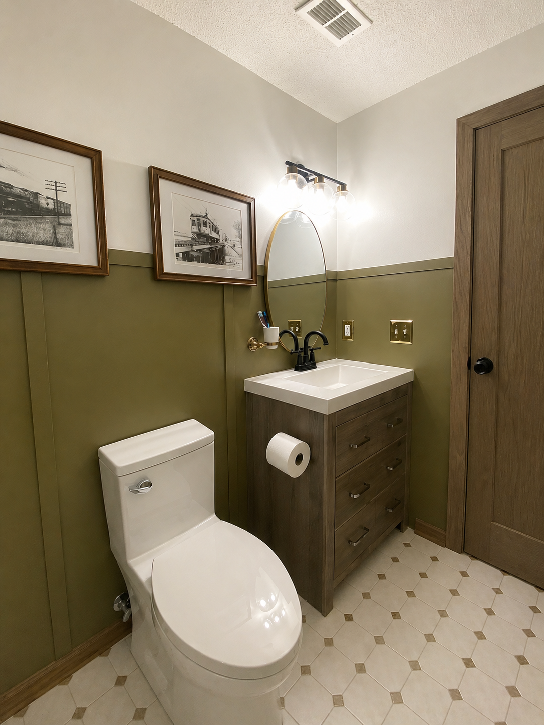 Small bathroom with a toilet, a wooden vanity with a sink, an oval mirror, wall-mounted light fixture, framed black-and-white train photos, green wainscoting, a towel roll, and light-colored tiled floor.