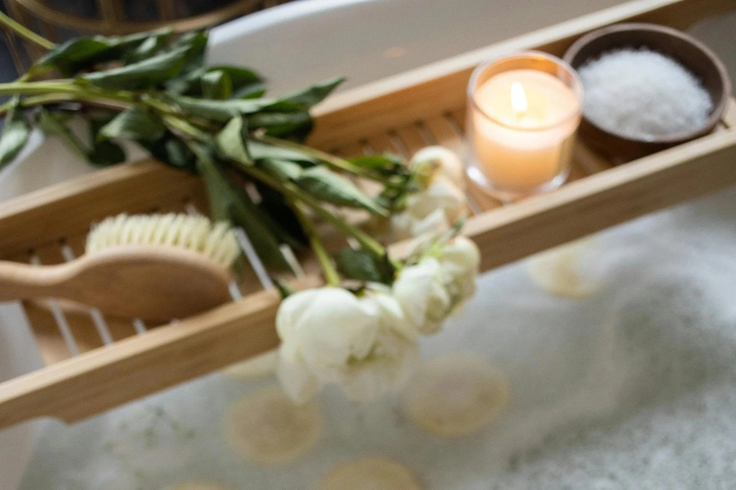 A wooden tray holding a lit candle, a bowl of coarse salt, and a bundle of green leaves and white flowers.