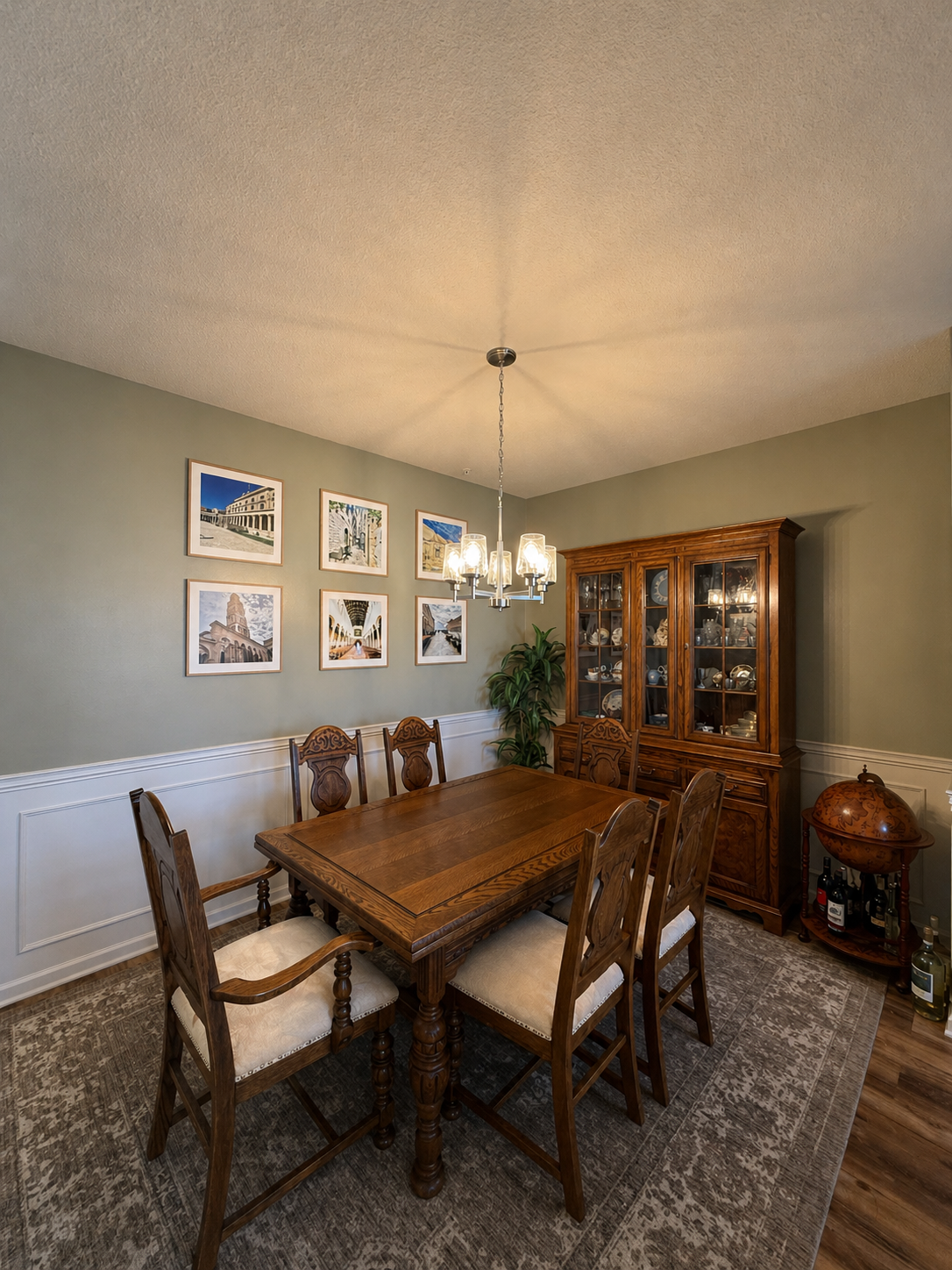 A dining room with a wooden table and six chairs, a china cabinet, framed photos on the wall, a globe, and bottles of alcohol on a small table.