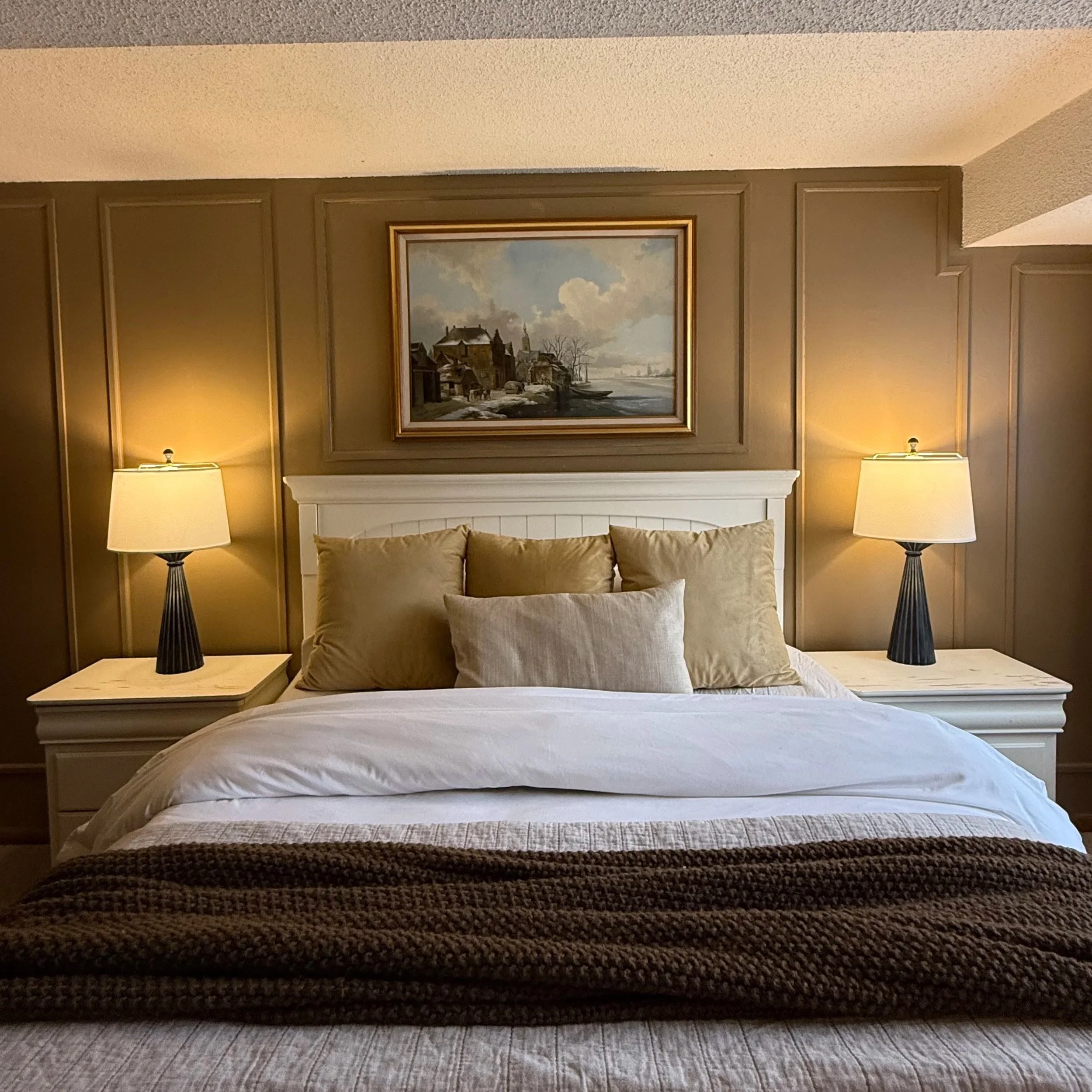 A bedroom scene with a bed, two lamps on nightstands, a painting above the headboard, and a textured blanket at the foot of the bed.