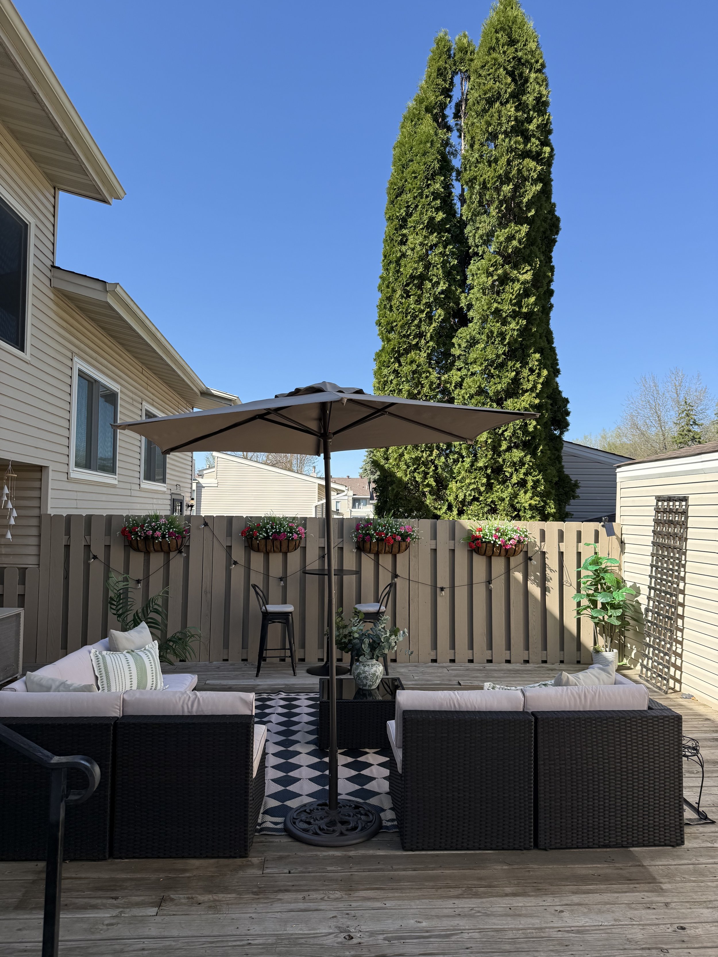 A backyard patio with black wicker furniture, white cushions, a black and white patterned rug, and a large beige patio umbrella. The yard is enclosed by a beige wooden fence decorated with potted pink and red flowers. There is a tall evergreen tree and the neighboring houses are visible against a clear blue sky.