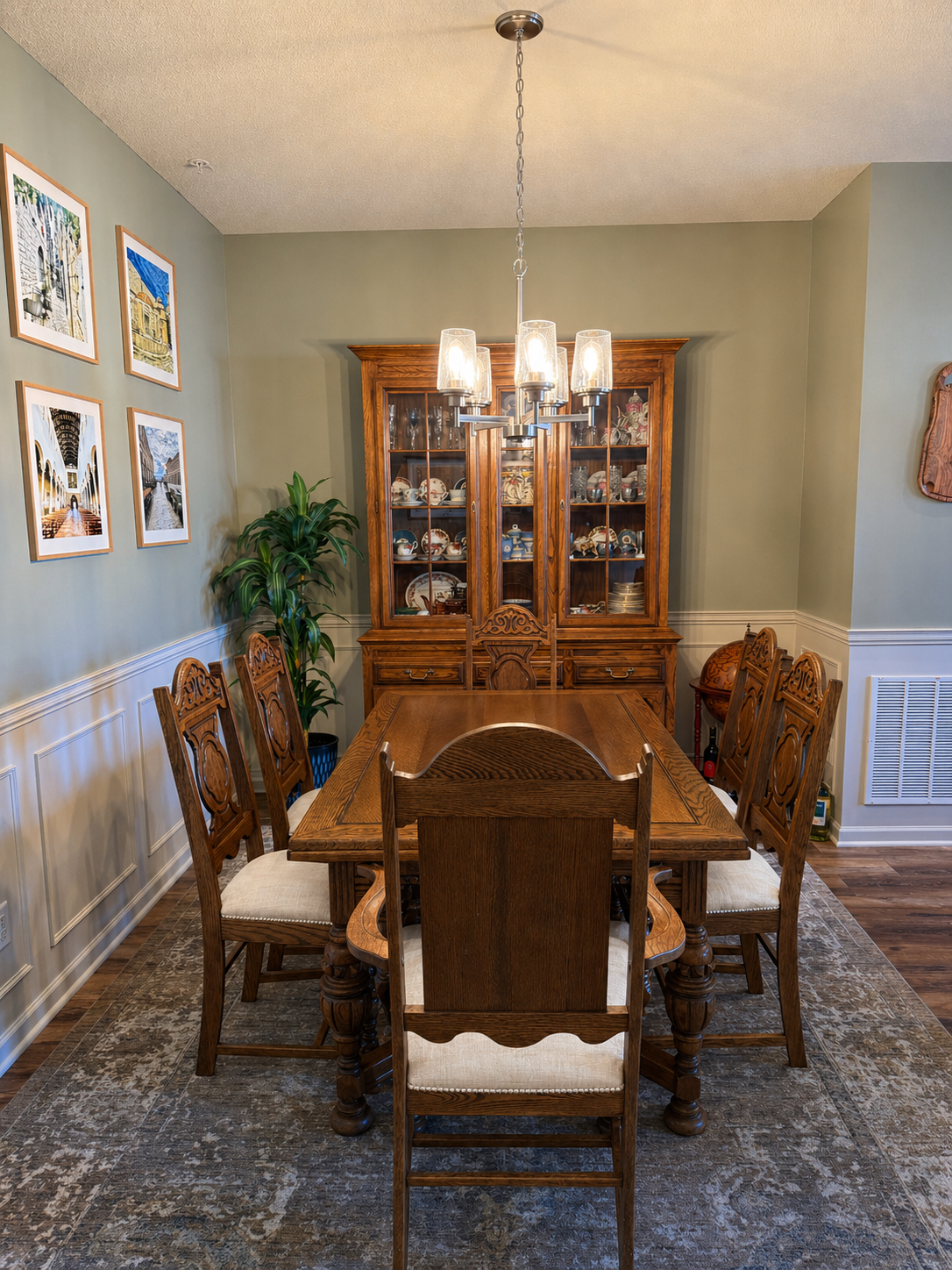 A dining room with a wooden dining table and six matching chairs with beige cushions. A wooden china cabinet filled with dishes, a potted plant, framed photos on the wall, and a chandelier hanging above the table.