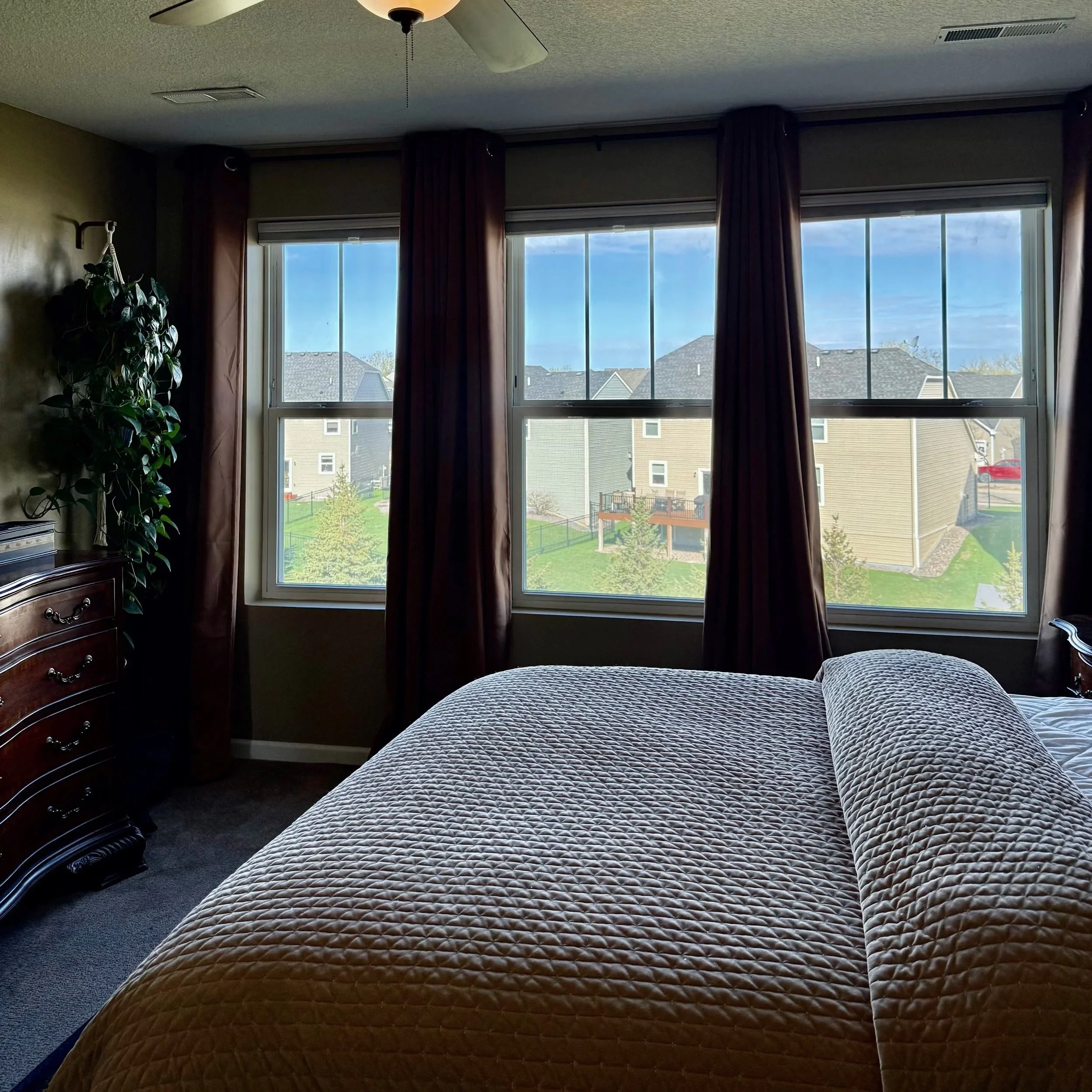 View of a bedroom with large window, closed curtains, and a view of neighboring houses and trees outside. Part of a bed with a quilted bedspread is visible in the foreground.