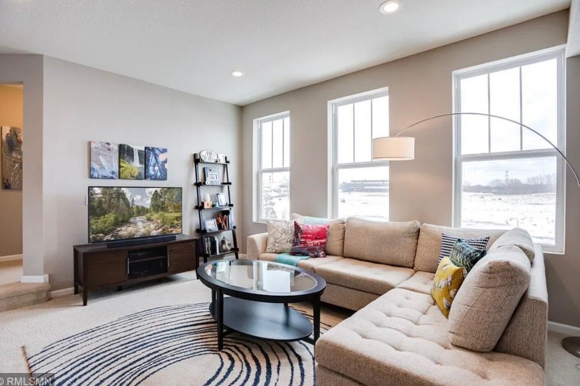 Living room with a large beige sectional sofa, floor lamp, black coffee table, flat-screen TV, black bookshelf, and three windows showing an outdoor snowy landscape.