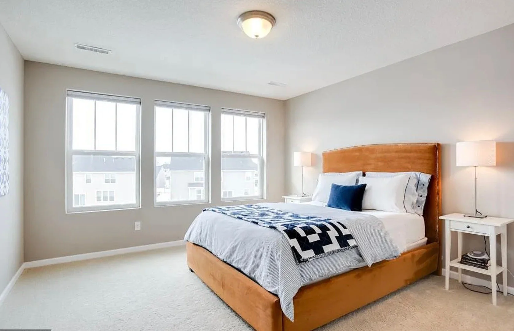 A bedroom with a wooden bed frame, white bedding, a blue pillow, and a black-and-white quilt. Two white bedside tables with lamps and books. Three large windows letting in natural light, with beige walls and carpet.