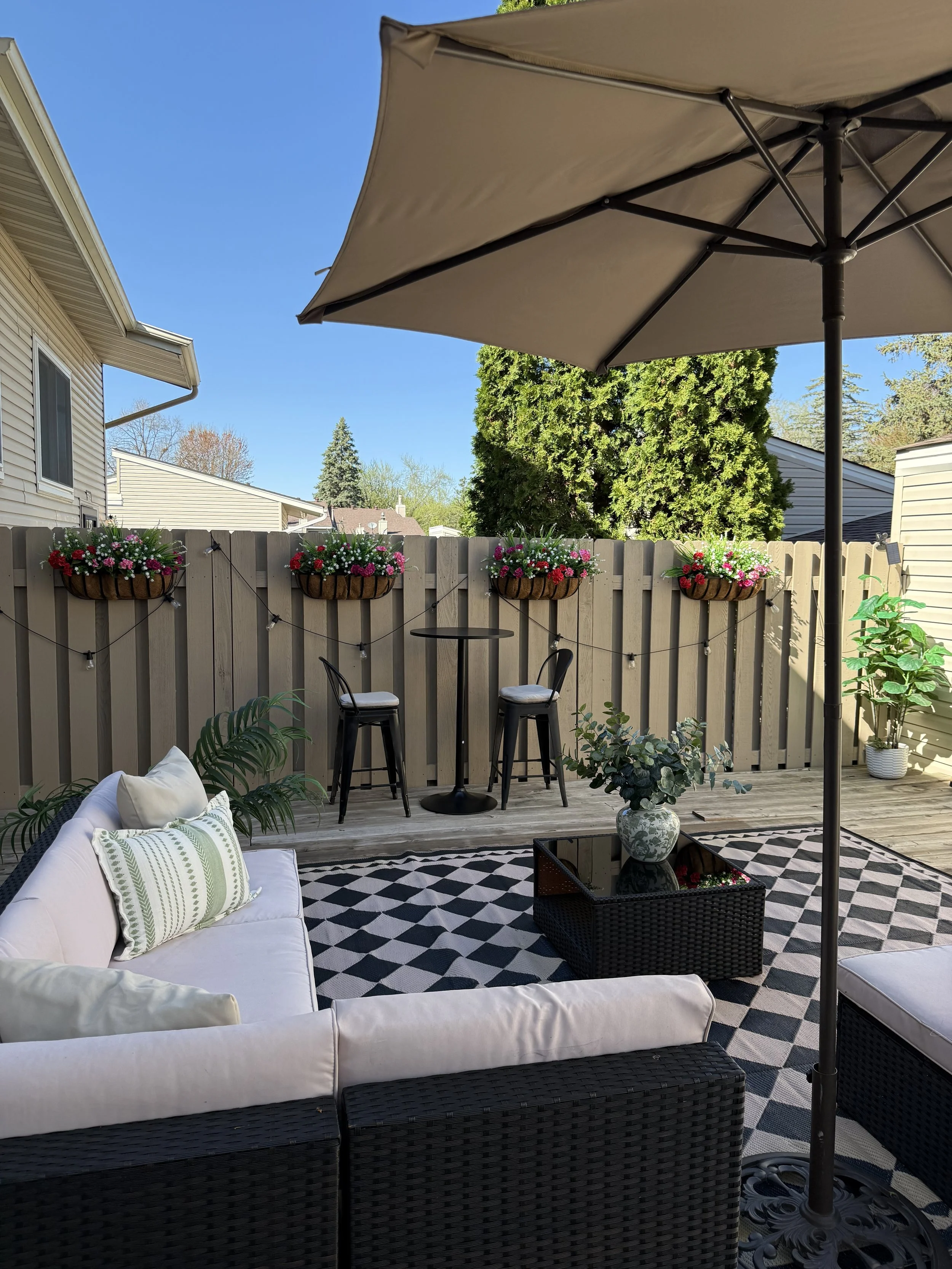 A cozy outdoor patio with a white sofa with decorative pillows, a black wicker coffee table with a potted plant, string lights, a large beige patio umbrella, and a wooden fence with flower baskets under a clear blue sky.