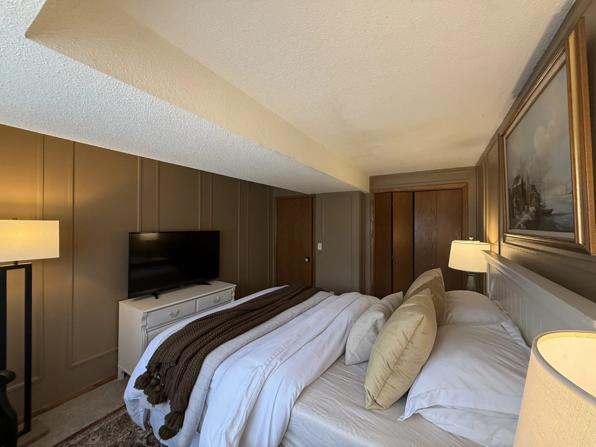 A bedroom with beige walls and a textured ceiling. There is a bed with white bedding and beige pillows. A white nightstand with drawers is beside the bed, with a television on top. Two table lamps are turned on, illuminating the room. Framed artwork hangs on the wall above the bed.