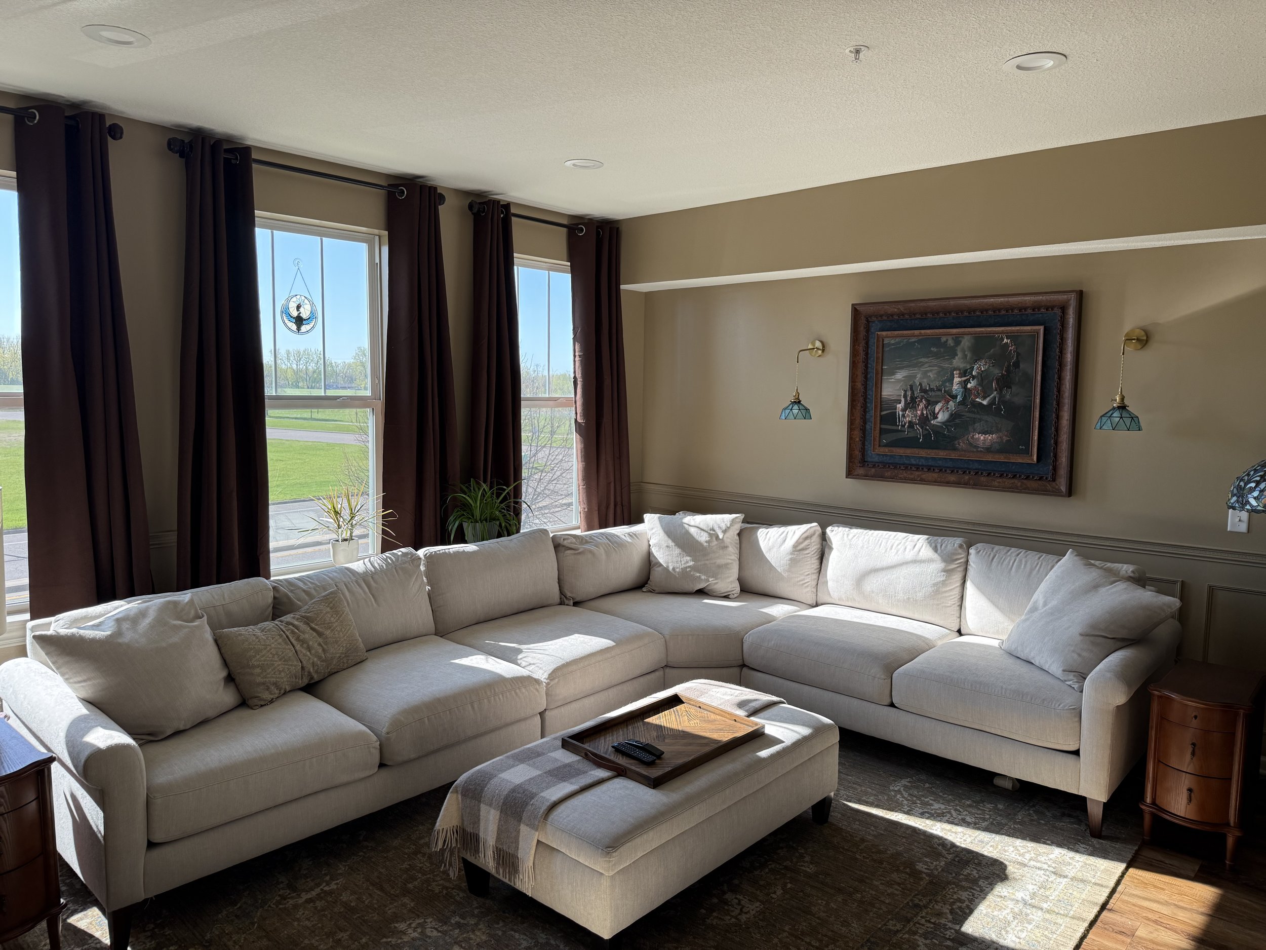 Living room with sunlight coming through large windows with brown curtains. There is a white sectional sofa with pillows, a small ottoman with a plaid blanket, a wooden tray with a remote on it, and framed artwork above the sofa. Small plants are on the window sill, and the room has beige walls and a dark carpeted floor.