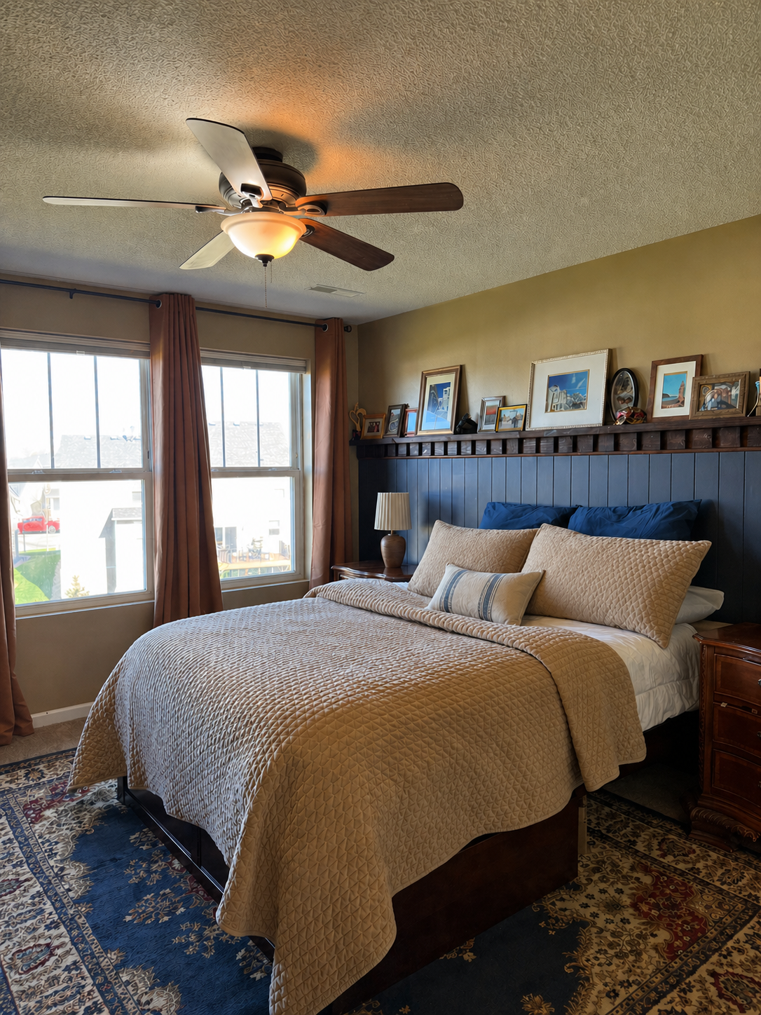 A cozy bedroom with a bed dressed in tan and blue pillows, a wooden nightstand with a lamp, a picture ledge with framed photos, and windows with orange curtains allowing natural light in.
