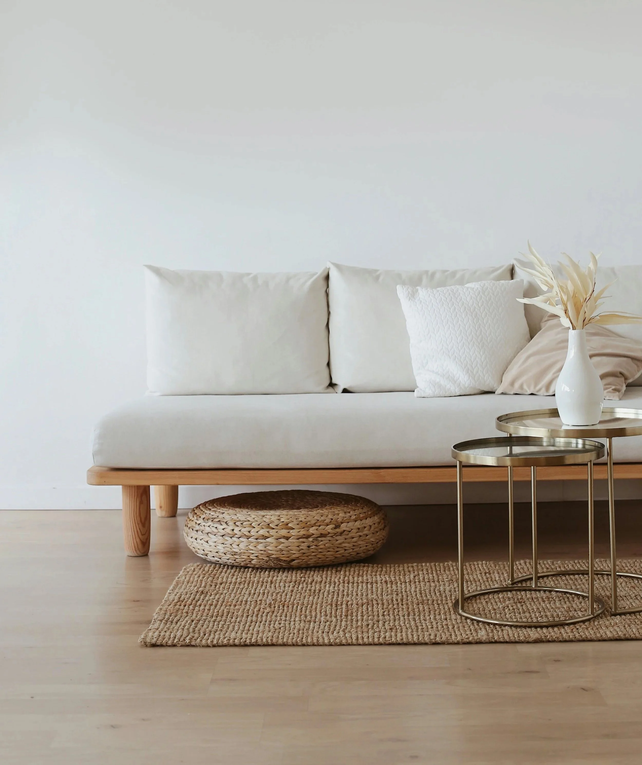 Minimalist living room with beige sofas, white textured pillows, a white vase with dried leaves, and side tables with woven rugs underneath on light wooden flooring.