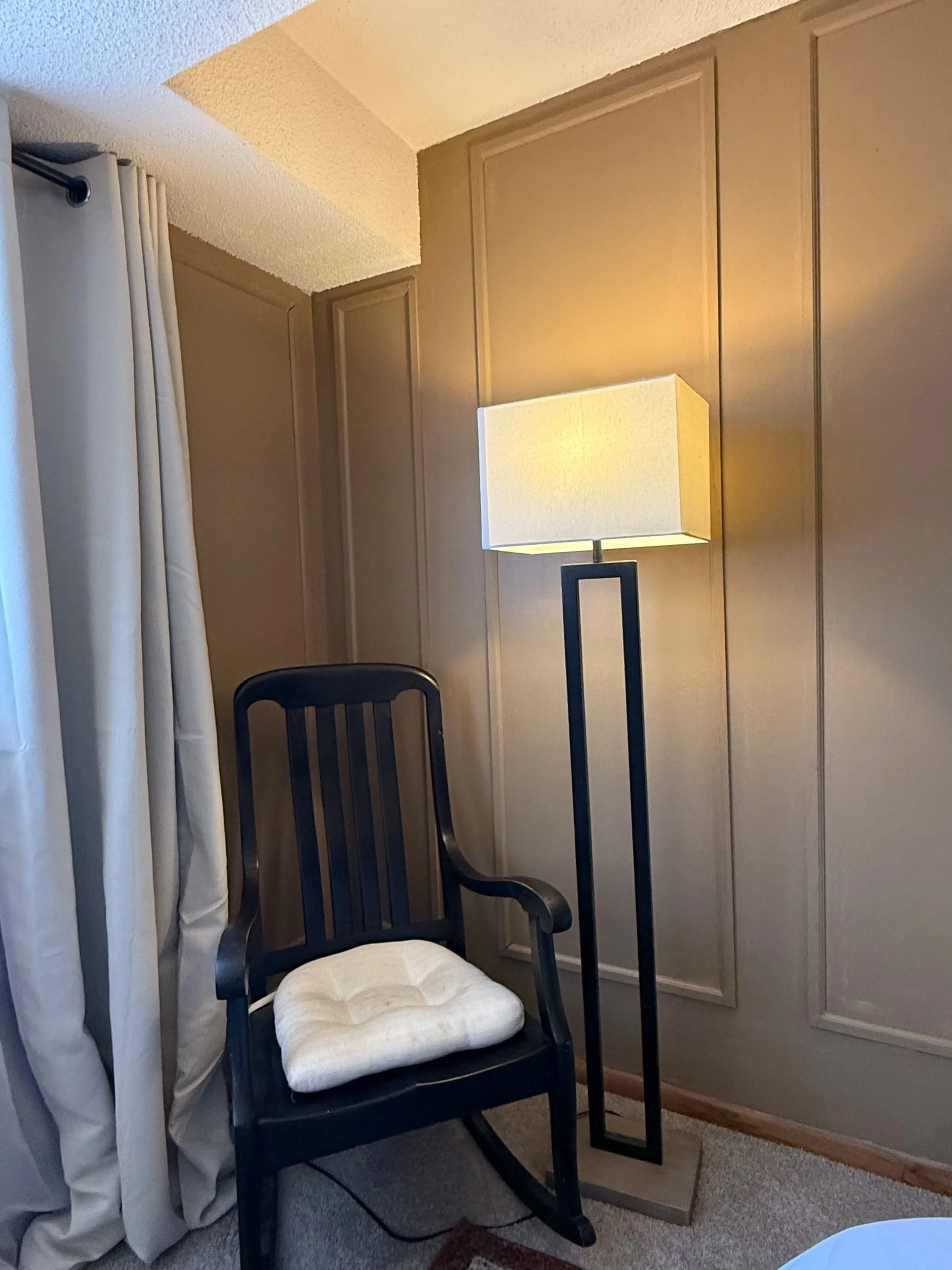 An interior room with beige walls and a beige ceiling. There is a black wooden chair with a white cushion. A white modern lamp with a rectangular shade is on a black stand. A beige curtain is hanging on the left side of the image.