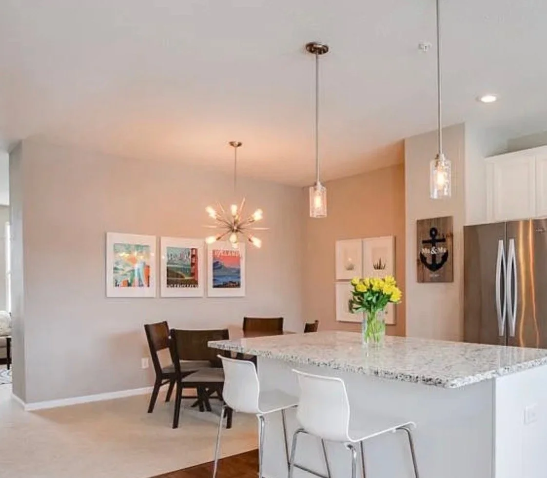 Modern kitchen and dining area with a granite island, white and black chairs, pendant lights, and wall art, with a vase of yellow flowers on the island.