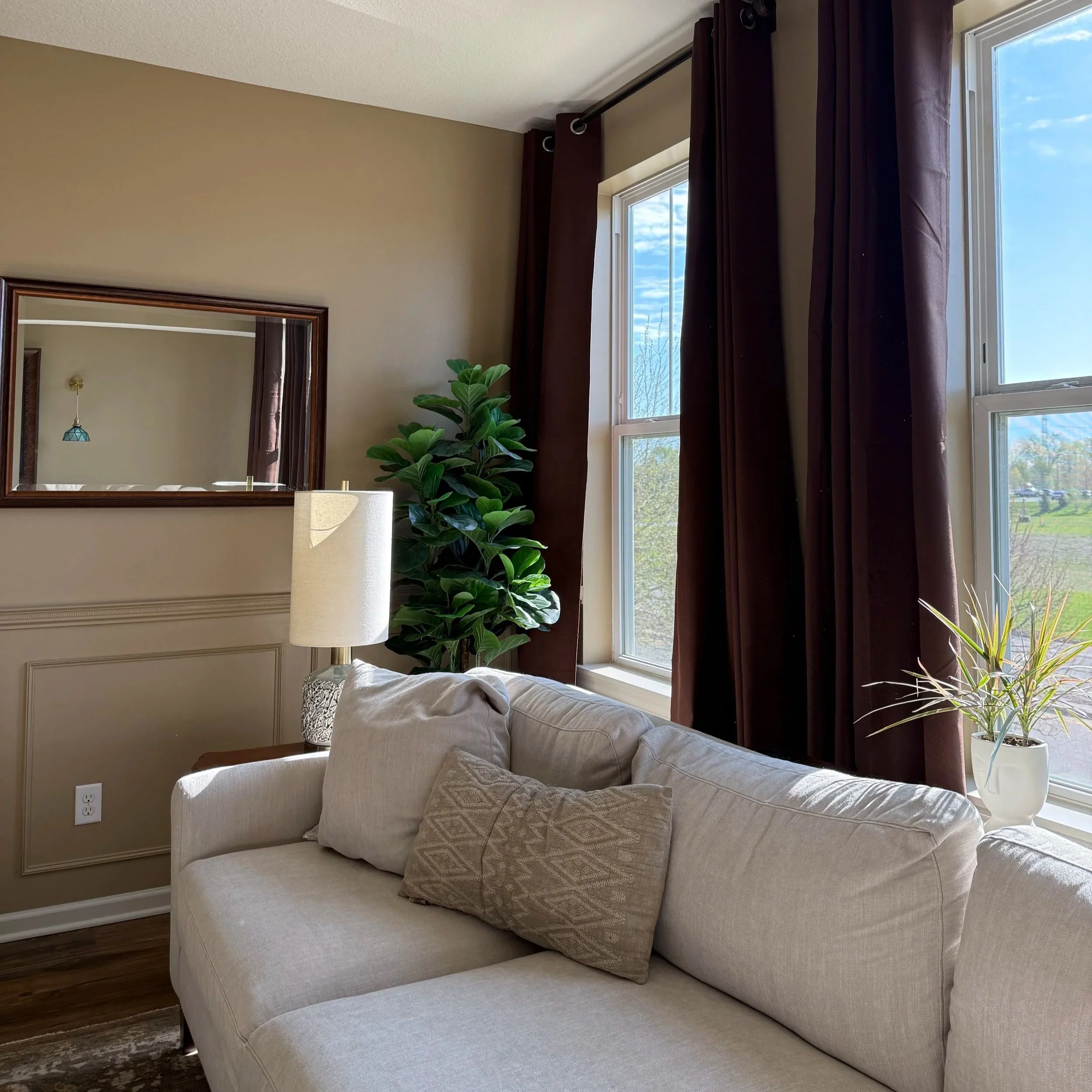 Living room with beige walls, a large beige sofa with throw pillows, a plant next to the window, and dark brown curtains.
