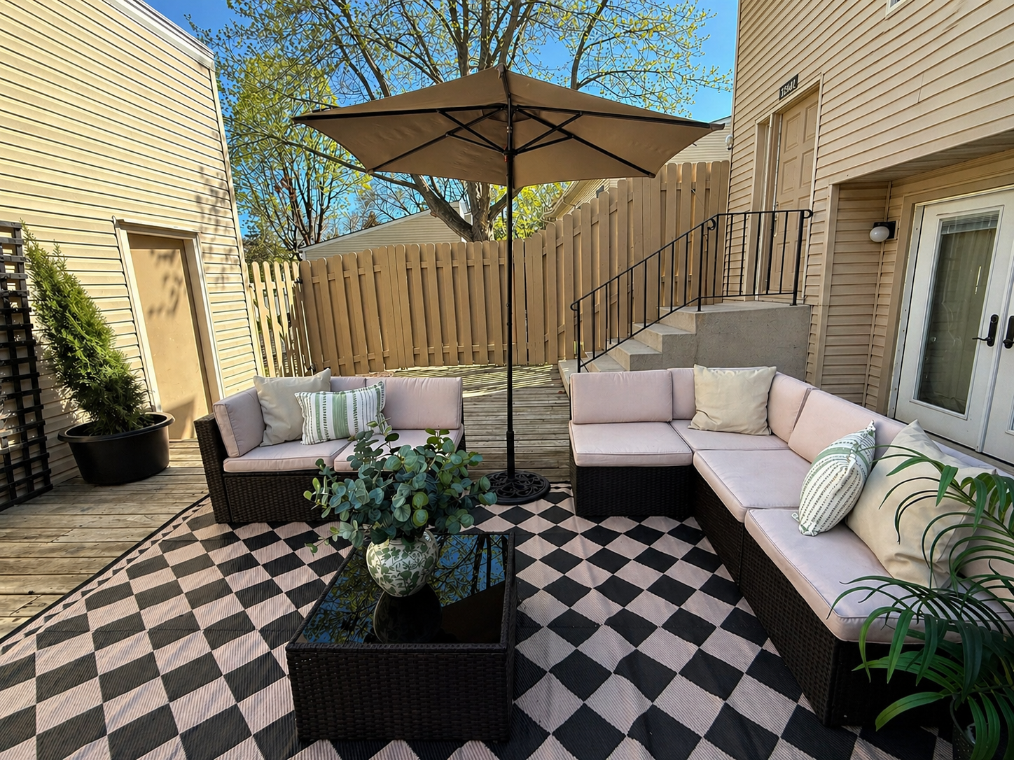 Backyard patio with woven outdoor sofa and chairs with cushions, centered with a black patio umbrella, underneath a large leafed plant in a vase, on a black and white checkered outdoor rug, adjacent to house with sliding door, potted plants, and a wooden fence behind.