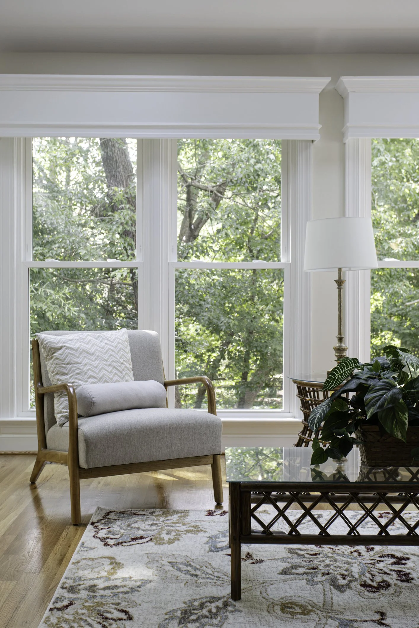 Sunlit living room with large window showing green trees outside, beige armchair with patterned pillow, glass-top table with woven base holding a potted plant, and a white floor lamp.