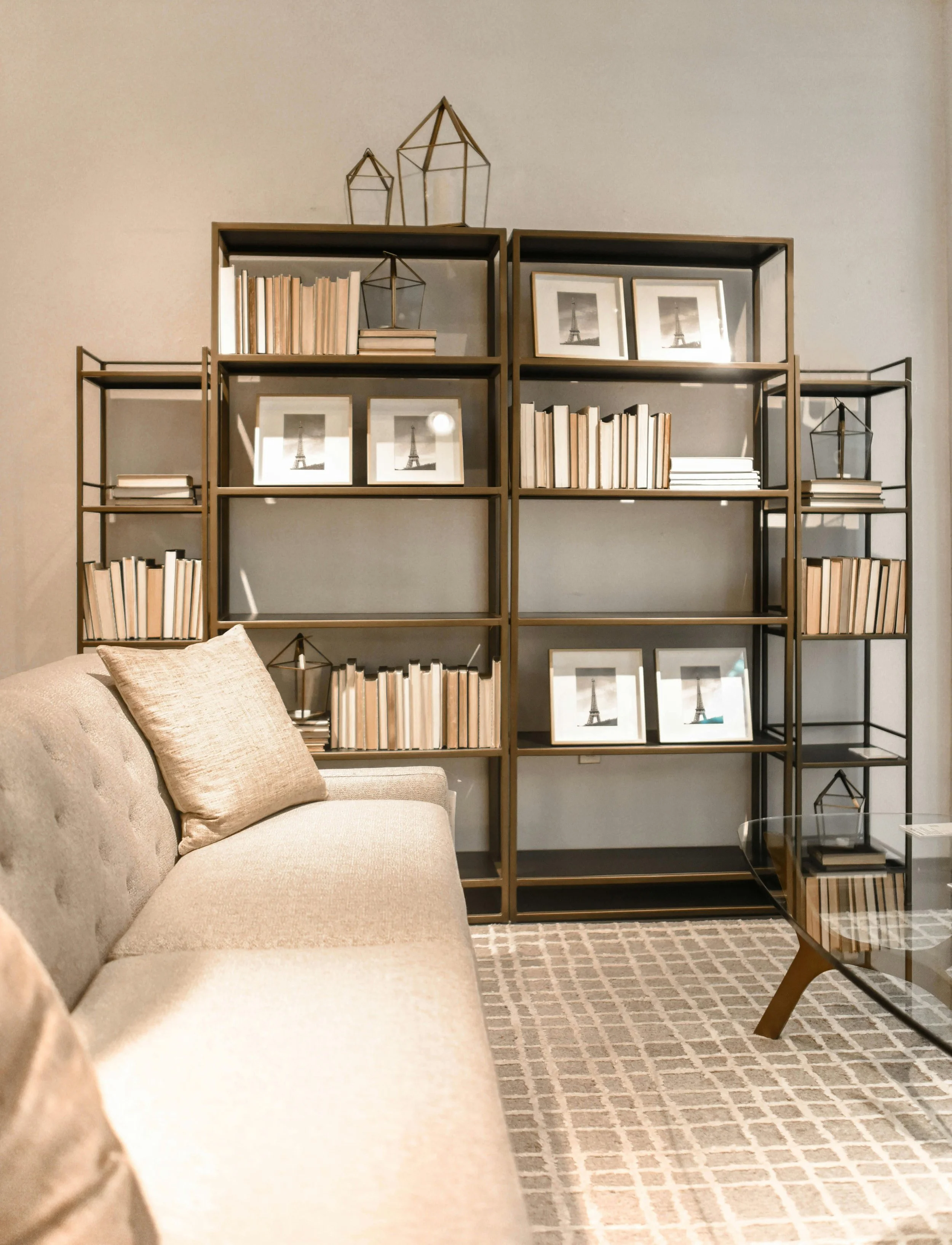 Living room wall with beige sofa, beige pillows, beige and brown patterned rug, black framed photographs of Eiffel Tower, bookshelf with books and decorative items, and a glass coffee table.